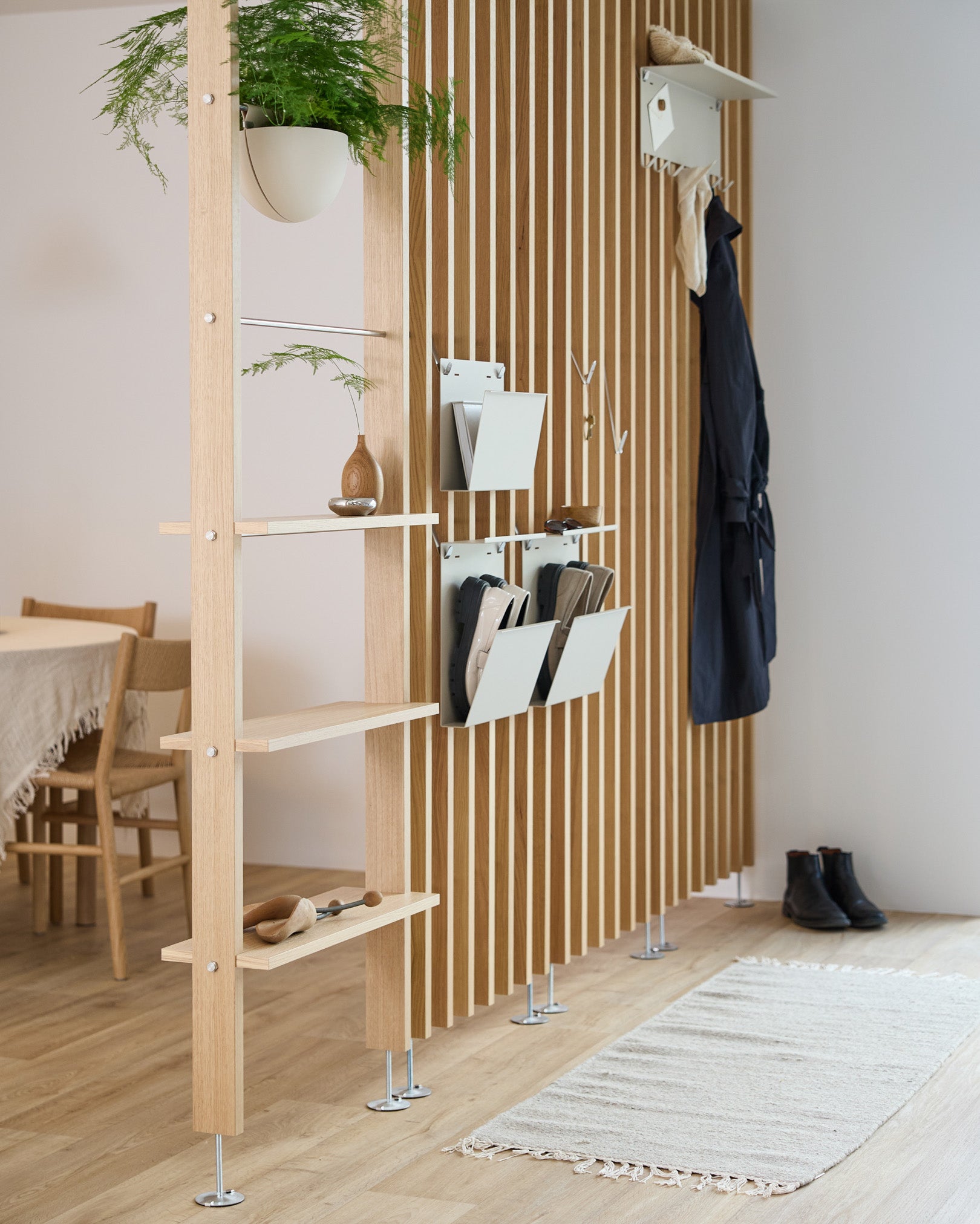 Nell Magazine Storage da WoodUpp em beige integrado num separador de ambientes com prateleiras em steel, caixas de arrumação e ganchos para casacos; ao fundo veem-se a zona de refeições e um tapete.