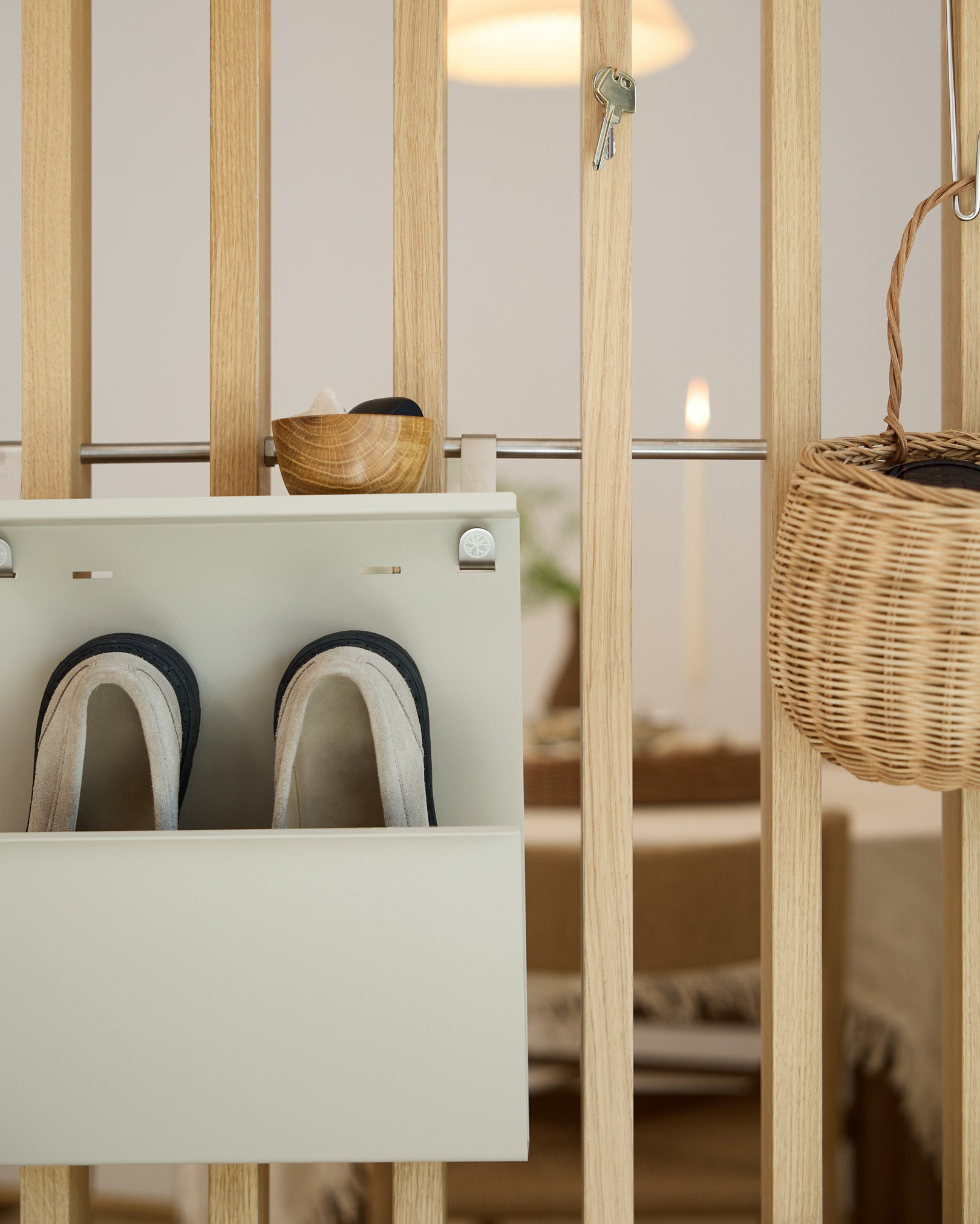 O Nell Beige Shoe Storage da WoodUpp apresenta chinelos, uma taça e um cesto de vime sobre um divisor de ambiente em madeira; ao fundo vê-se a zona de refeições.