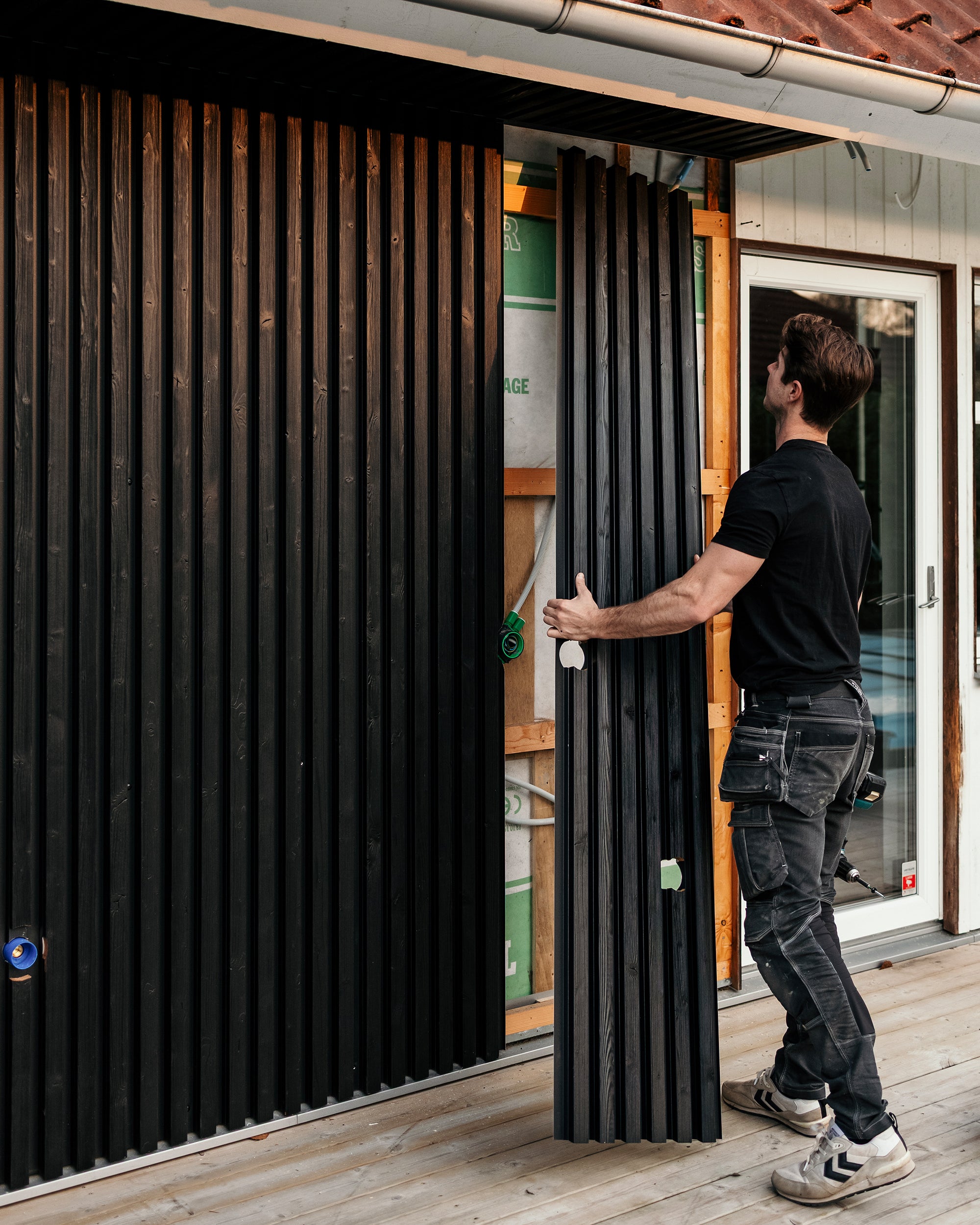 Um homem instala painéis de fachada Aluwood pretos em posição vertical na parede exterior de uma casa enquanto está em pé num deck recém-construído.