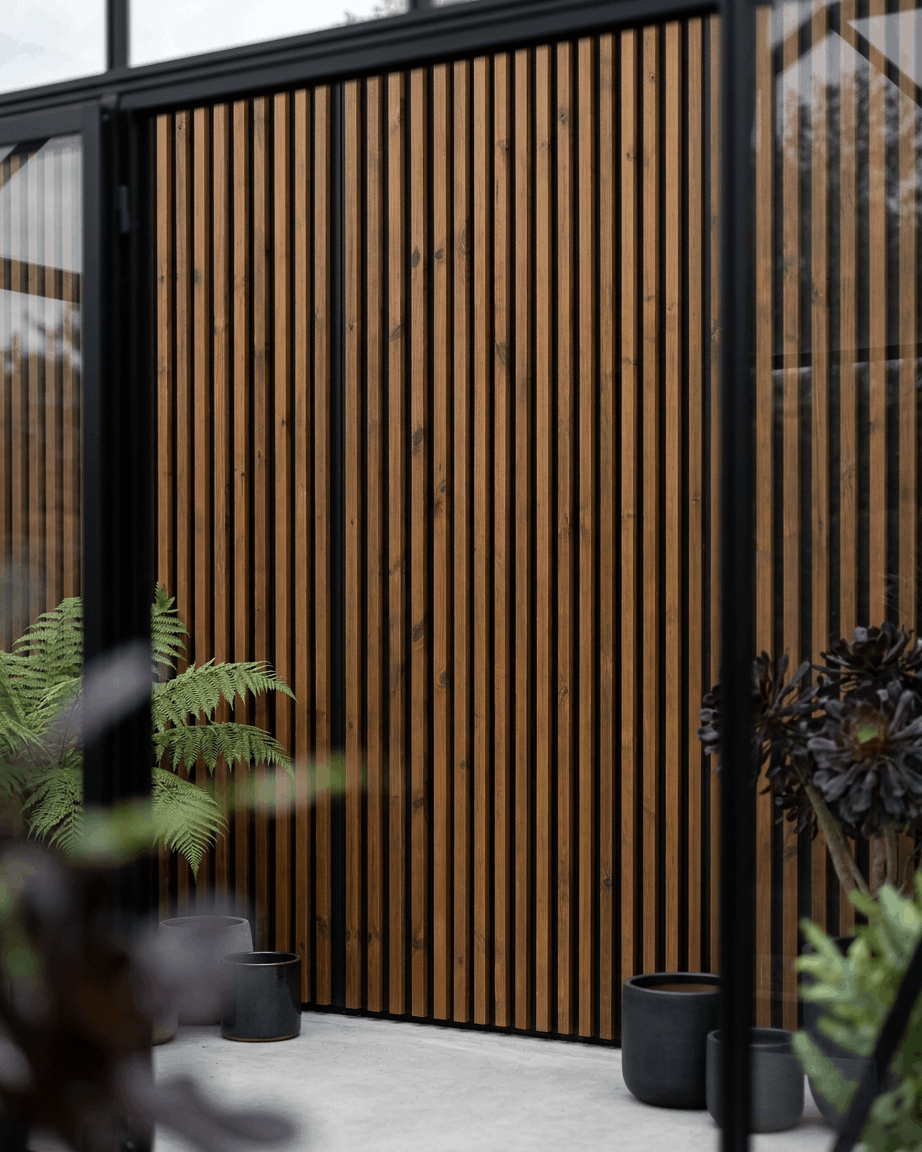 Painel de fachada exterior WoodUpp Aluwood em brown oleado, combinado com floreiras black e plantas verdes, criando um ambiente de pátio nórdico minimalista.
