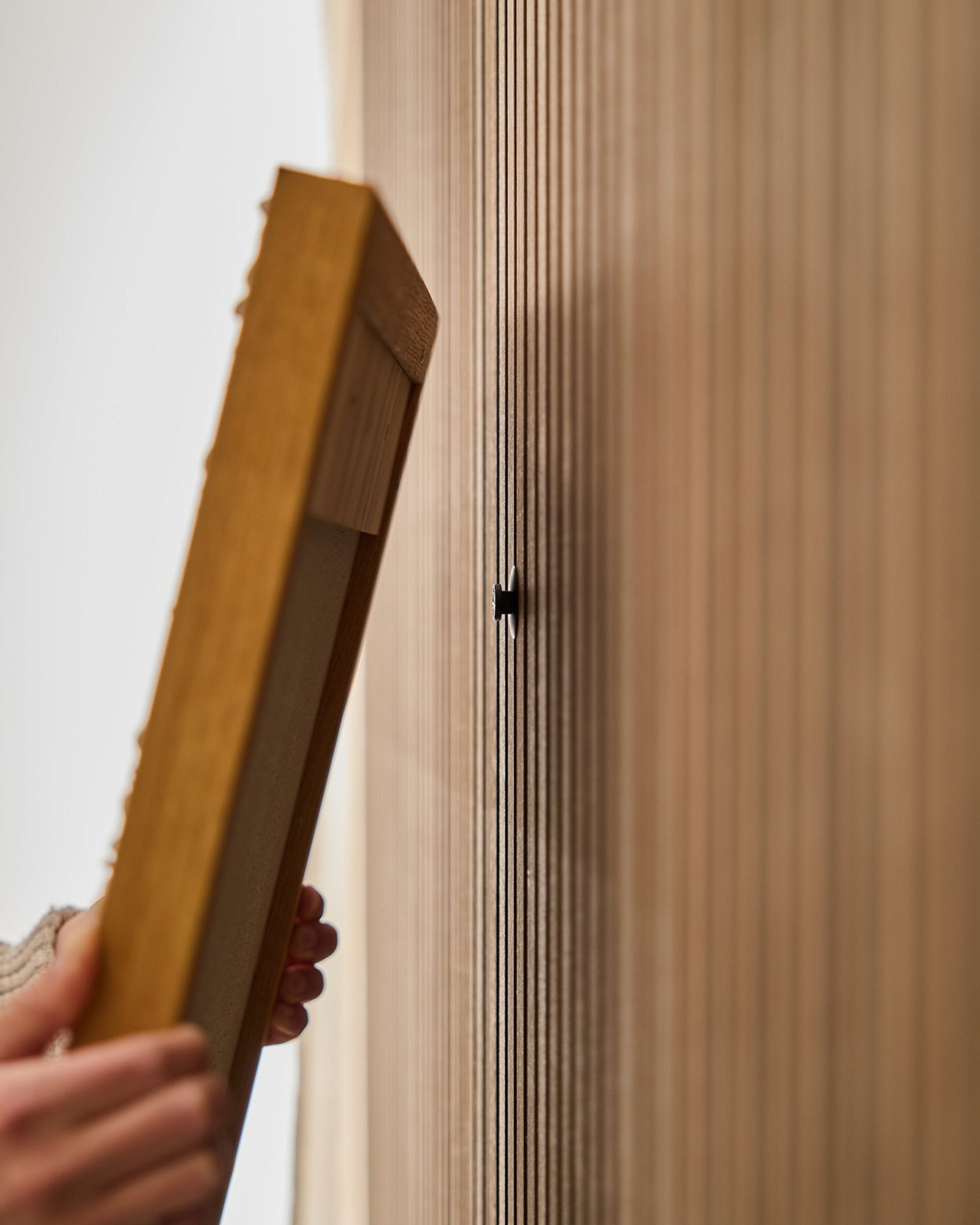 Uma pessoa usa o conjunto WoodUpp Dupps | Original para montar uma estrutura de painel acústico em madeira, focando-se na fixação de um gancho small black junto às ripas vertical.