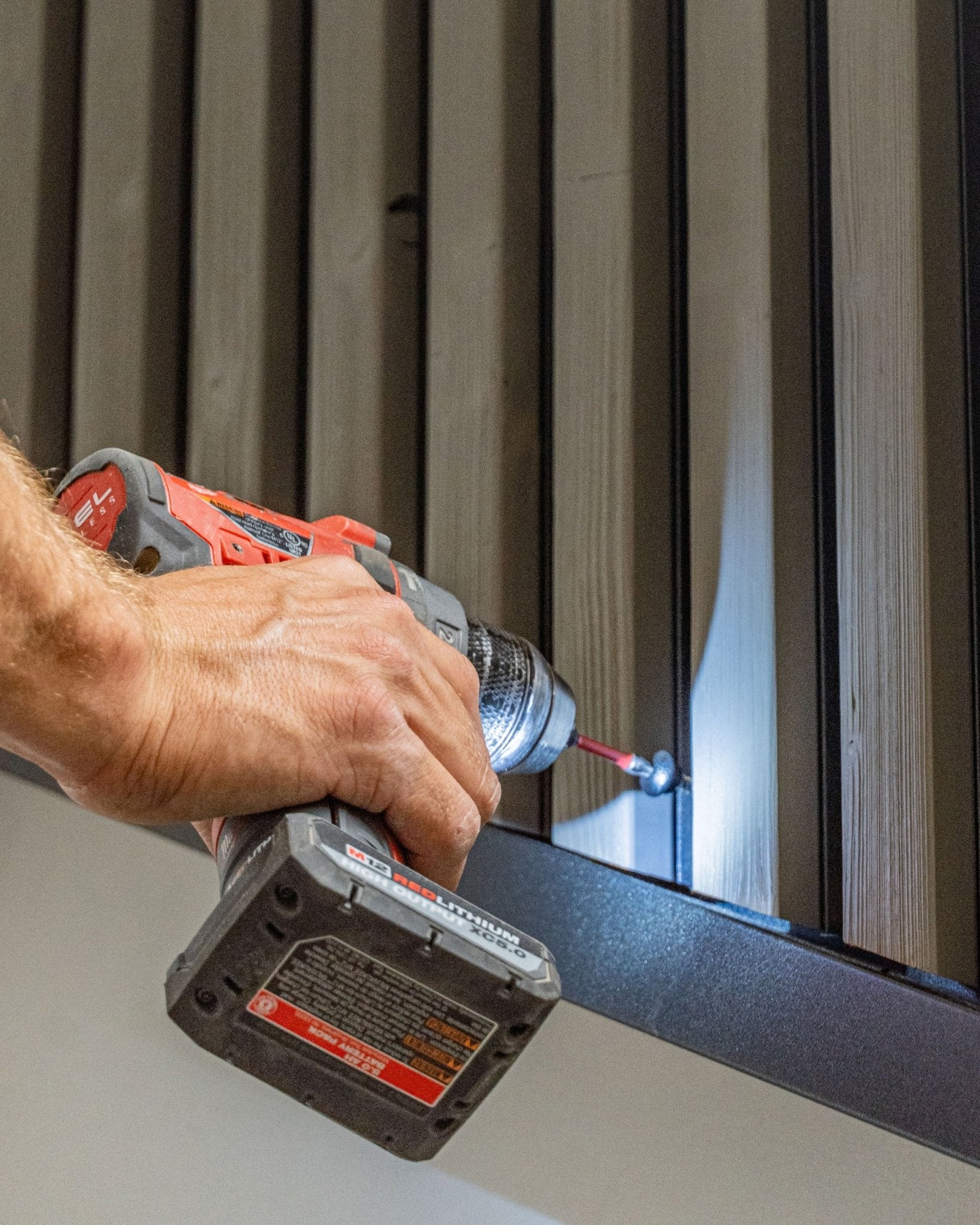Uma pessoa fixa um parafuso frontal WoodUpp black (4.8x35, anilha Ø9.5) num painel de fachada vertical Aluwood com um berbequim sem fios.