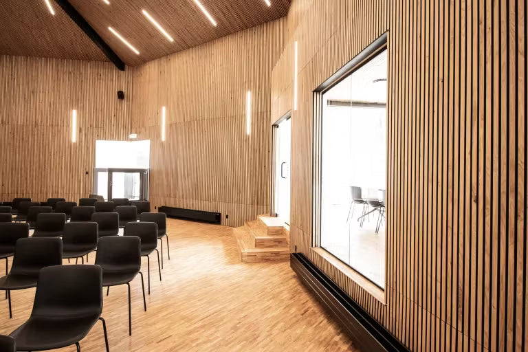Sala de conferência espaçosa com painéis de madeira acústica Akupanel nas paredes e no teto, fileiras de cadeiras pretas, janelas grandes e luminárias verticais.