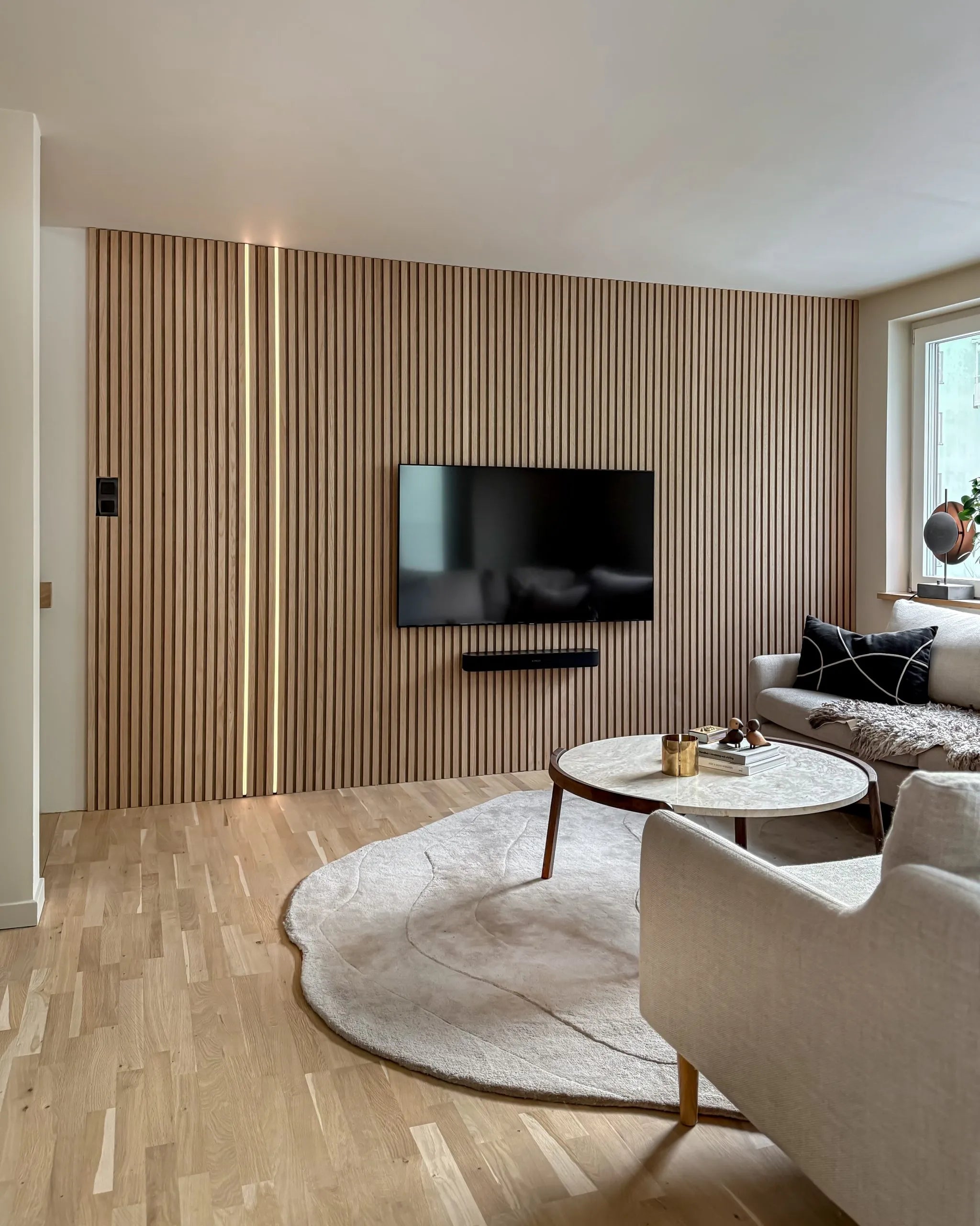 Sala de estar moderna com uma parede nórdica de ripas de madeira acústica Akupanel, TV montada, mesa de centro redonda, sofá creme, tapete grande sobre piso de madeira clara.