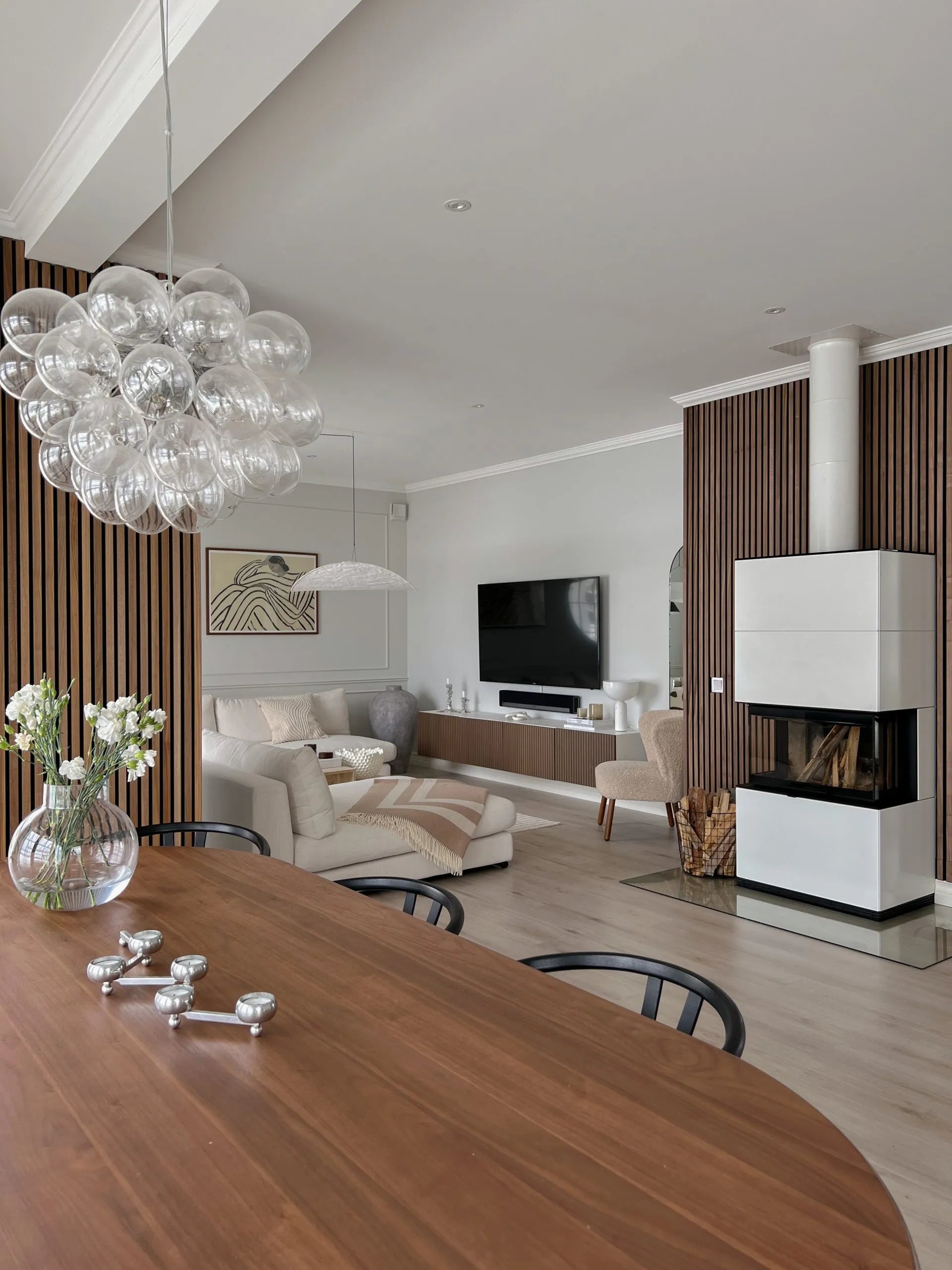 Moderna área de estar e jantar com painéis acústicos de madeira nórdica, assentos aconchegantes, TV, lareira elegante com lenha empilhada e um lustre de bolhas de vidro.