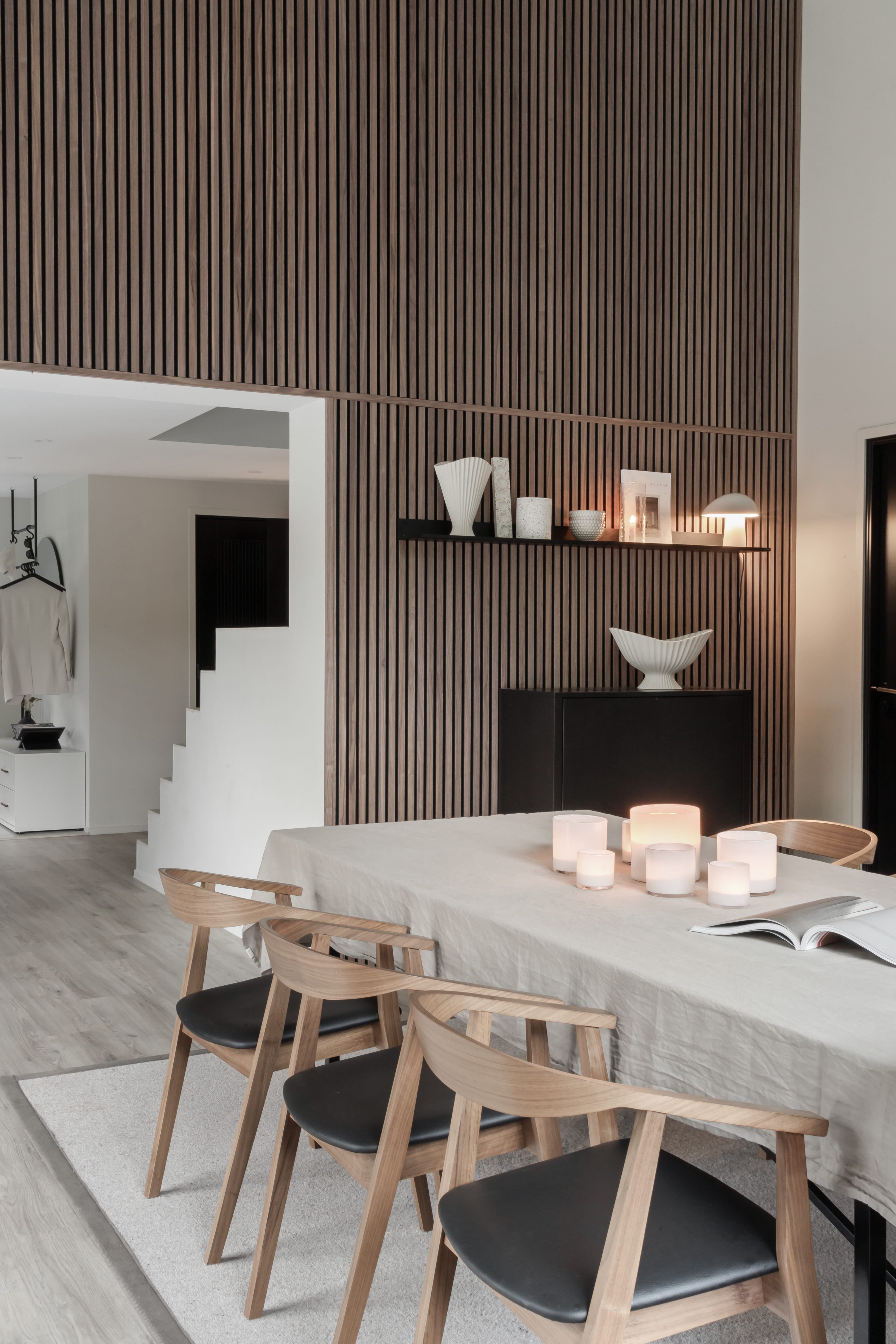 Sala de jantar moderna com uma mesa de madeira clara, quatro cadeiras, velas e decoração minimalista. Parede vertical de ripas de madeira acústica Akupanel e escada aberta à vista.