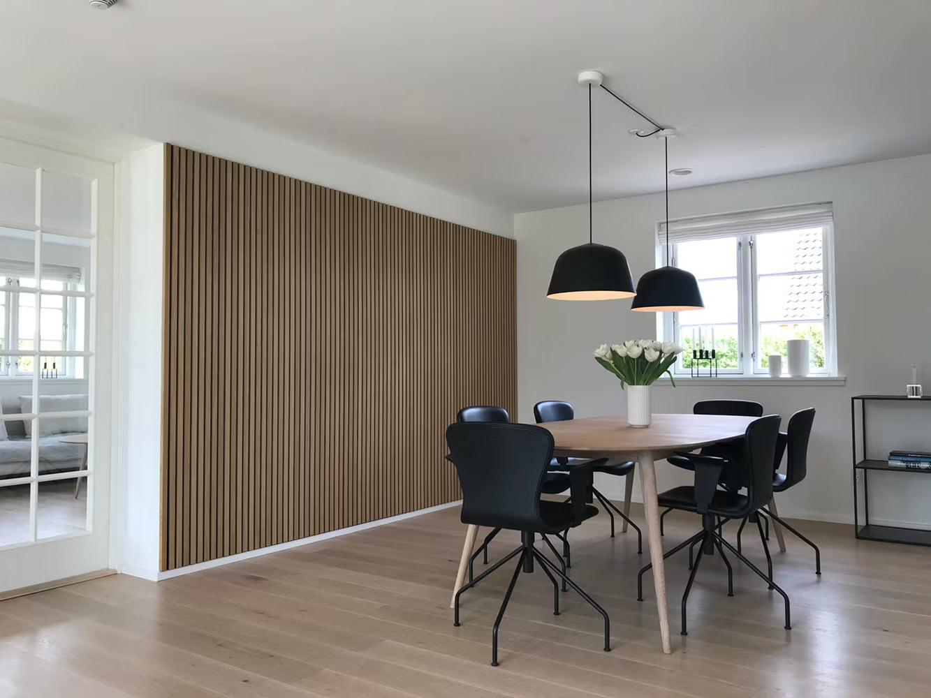 Sala de jantar moderna com piso de madeira, parede de ripas acústicas Akupanel, cadeiras pretas, mesa de madeira, luminárias pendentes e um centro de mesa floral branco.