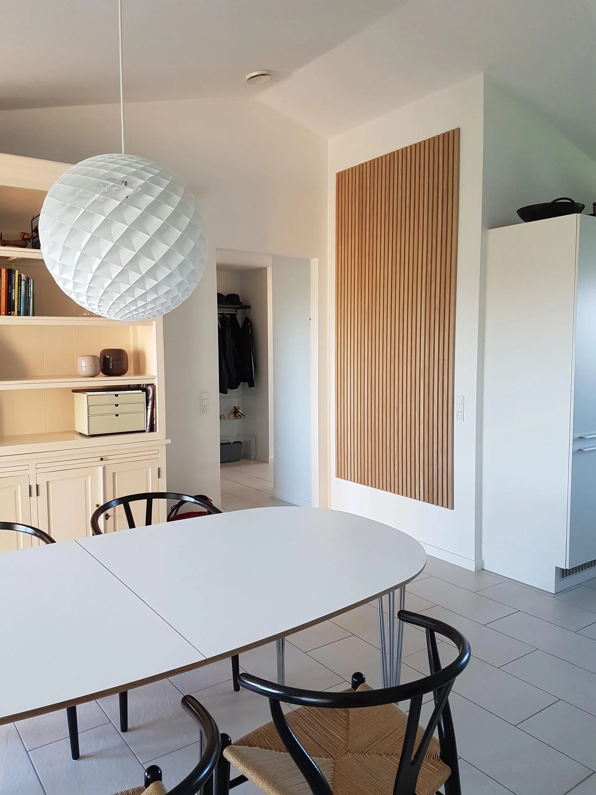 Sala de jantar moderna com uma mesa oval branca, cadeiras pretas, luminária pendente grande e texturizada e armários acústicos Akupanel de madeira clara e elegante.