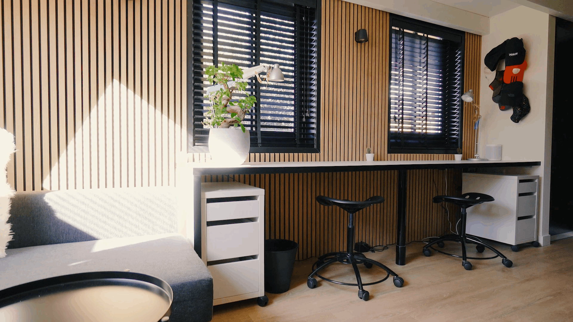 Escritório moderno com painéis de madeira acústica nórdica nas paredes, estações de trabalho ergonômicas, banquetas pretas, um vaso de plantas e luz solar através de persianas pretas.