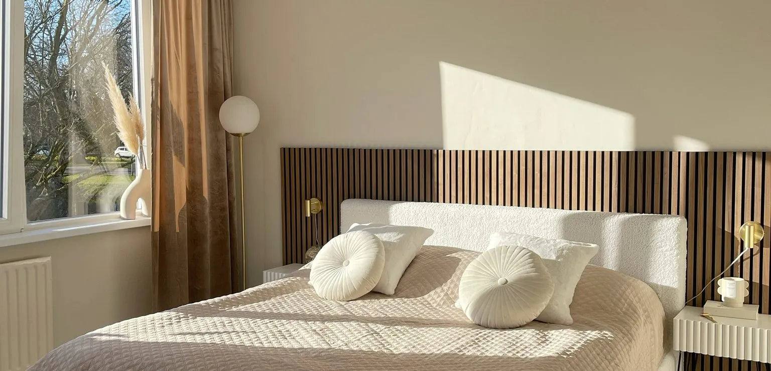 Quarto moderno iluminado pelo sol com uma cama acolchoada bege, travesseiros redondos, cabeceira de ripas de madeira Akupanel e decoração minimalista ao lado de uma grande janela com cortinas transparentes.