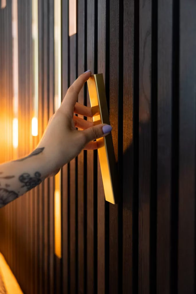 Uma mão tatuada com unhas roxas claras segura uma alça dourada em uma parede de painel de madeira acústica Akupanel escura e elegante, realçada por uma iluminação quente.
