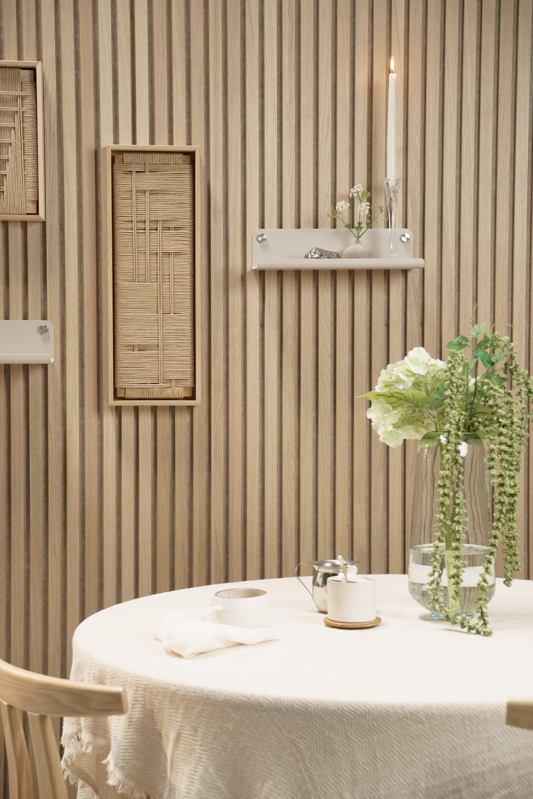 Uma mesa redonda com um pano branco, vaso floral, vela e decoração colocada em frente a uma parede de ripas acústicas Akupanel de madeira clara com detalhes em tecido e prateleiras.