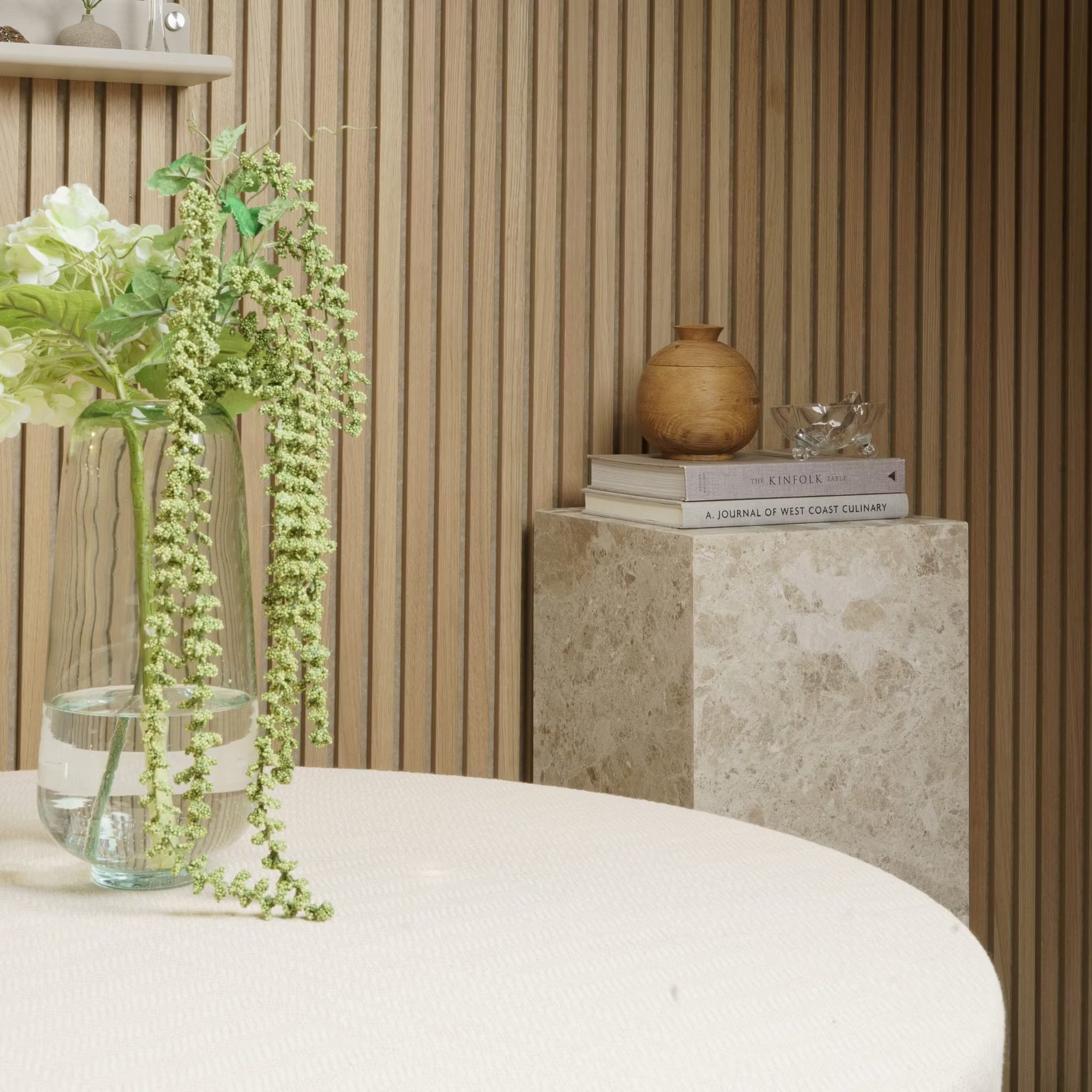 Um vaso com flores fica em uma mesa redonda; livros e cerâmicas descansam em um pedestal de mármore contra uma parede de painel de madeira acústica Akupanel.