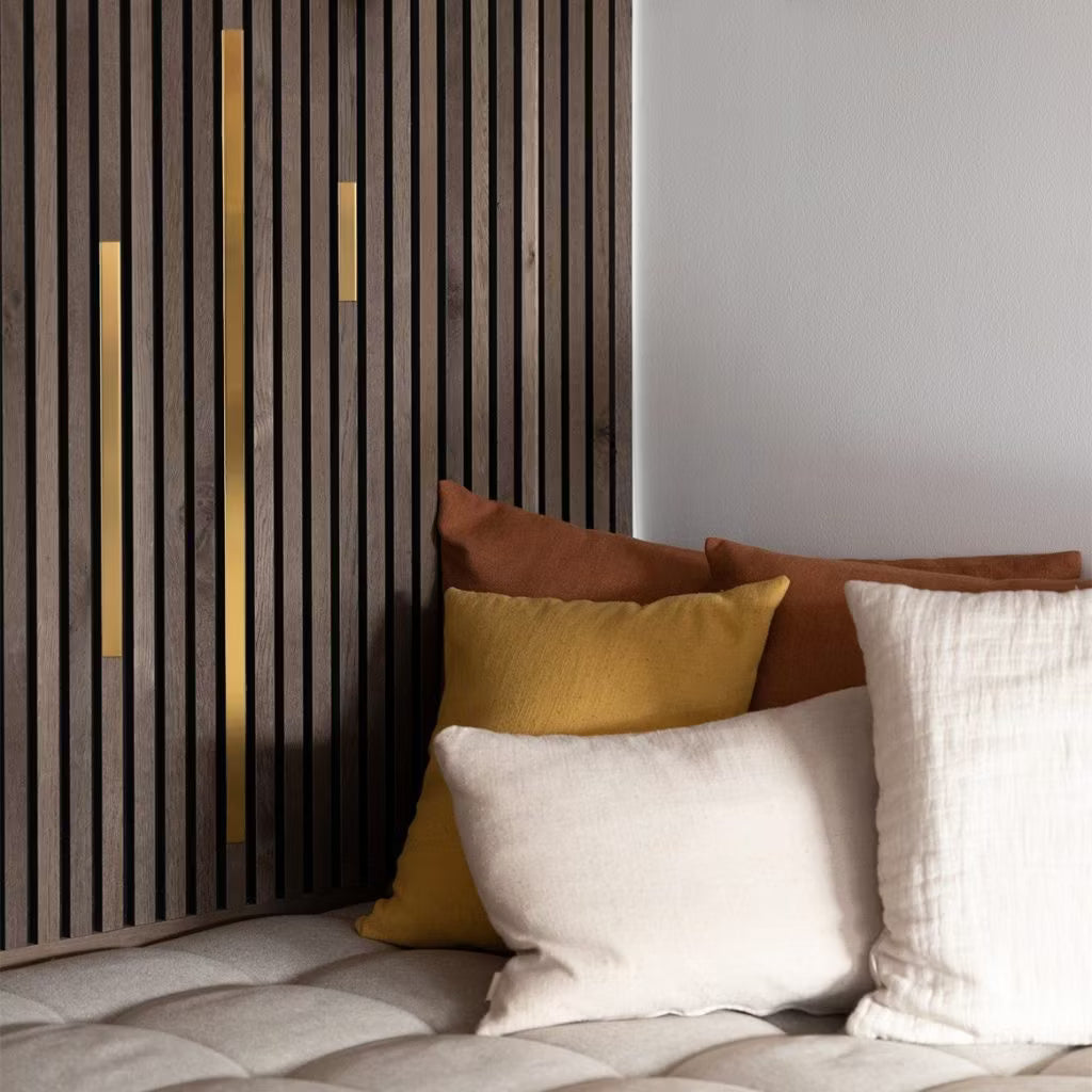 Almofadas em tons de creme, amarelo e marrom em um sofá bege com tufos, ao lado de um painel acústico moderno de parede de ripas com detalhes em dourado para um ambiente aconchegante e contemporâneo.