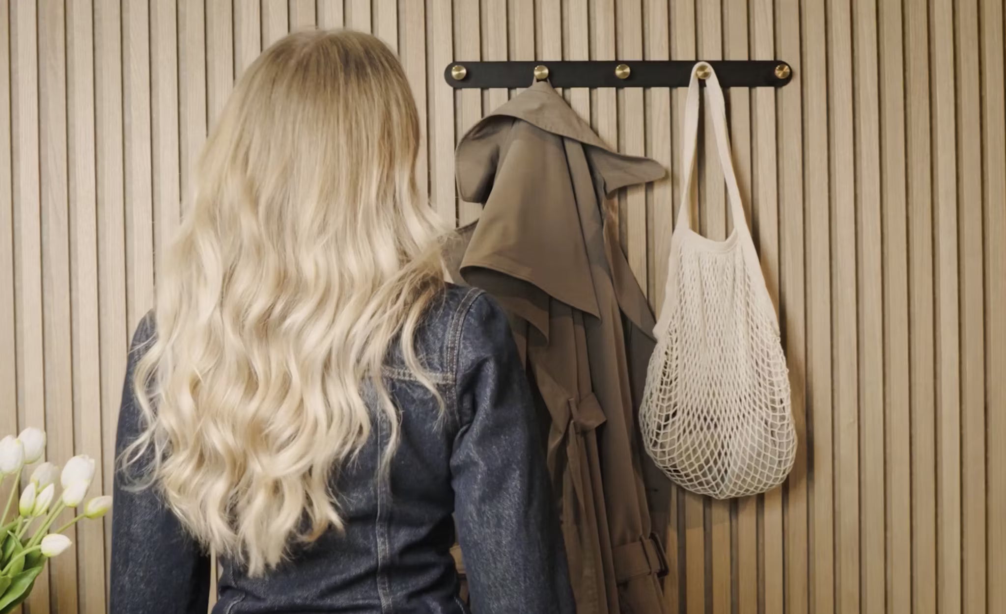 Uma mulher com longos cabelos loiros está diante de um cabide com um sobretudo, uma bolsa de rede branca e tulipas brancas frescas ao lado de um painel de madeira acústica Akupanel no canto.