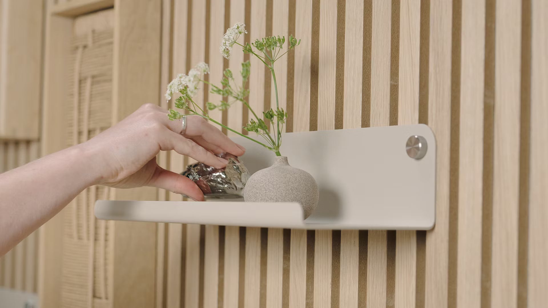 Uma mão coloca um pequeno objeto decorativo ao lado de um vaso de flores brancas em uma prateleira minimalista contra um painel de ripas de madeira acústica nórdica texturizada.