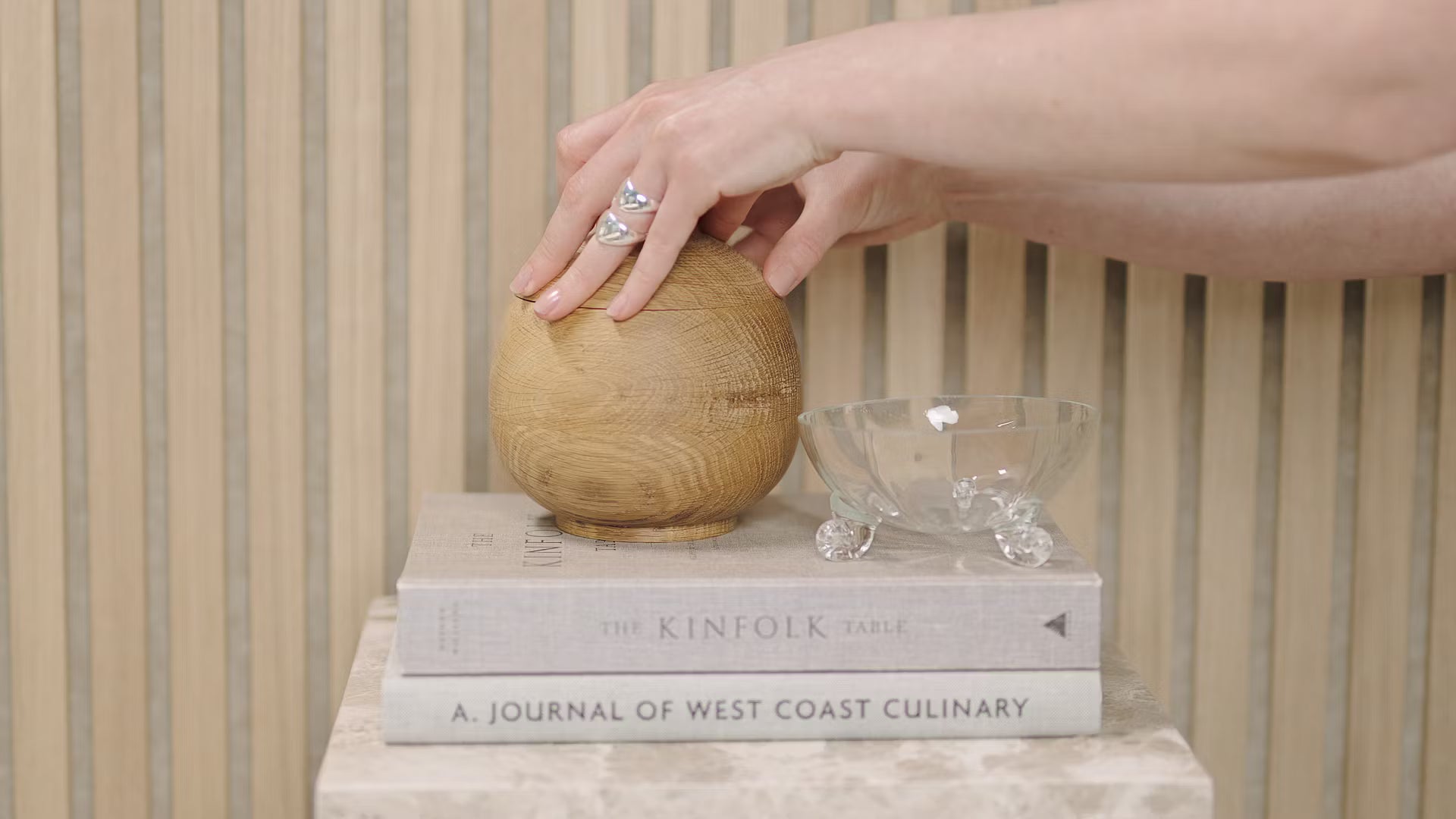 Uma mão toca uma esfera de madeira colocada sobre livros empilhados, incluindo Kinfolk Table e Journal of West Coast Culinary, ao lado de uma tigela de vidro em um painel acústico de ripas de madeira.