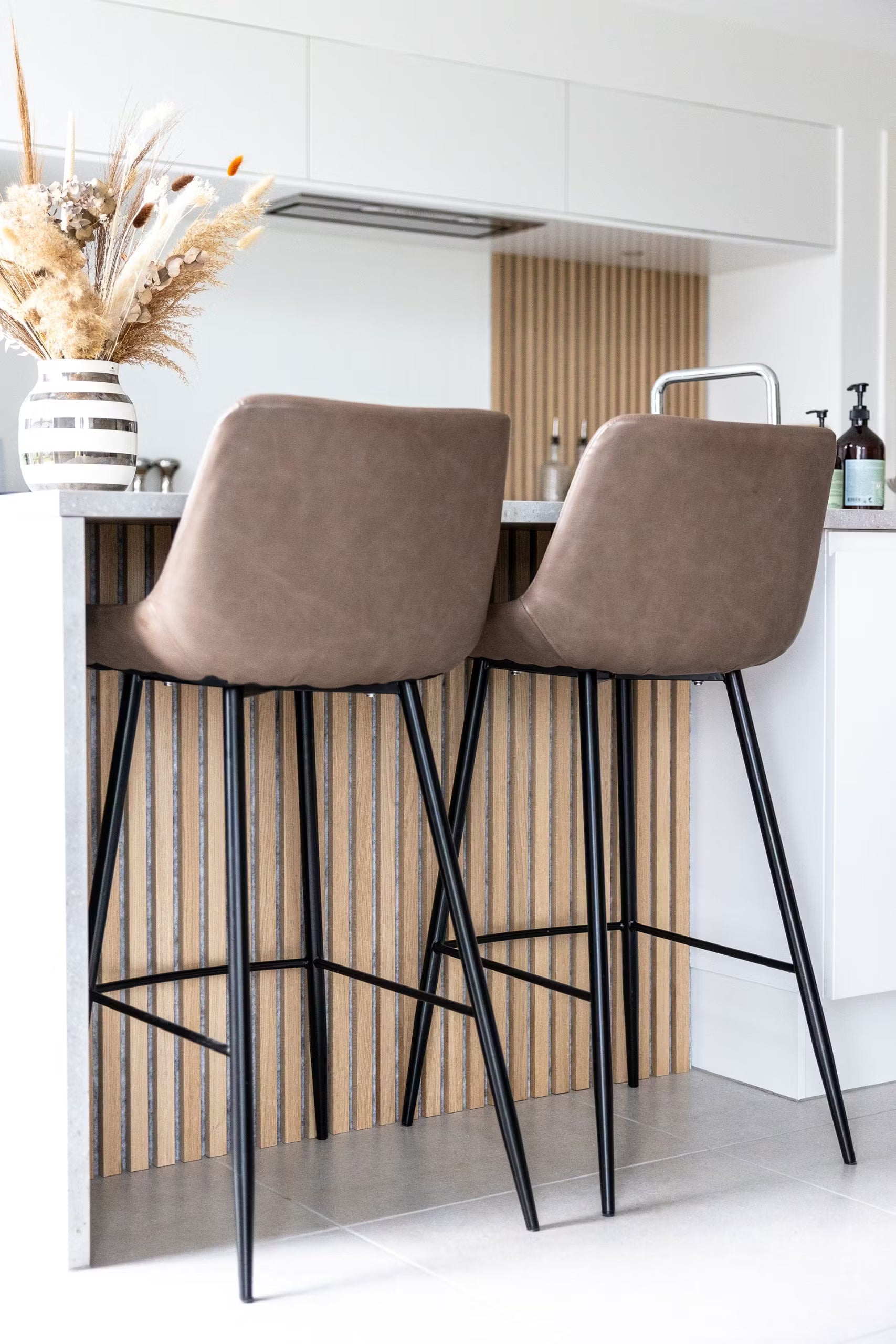Banquetas de bar marrons com pernas pretas em uma ilha de cozinha, com um painel de ripas de madeira acústica nórdica e um vaso com flores secas na bancada.