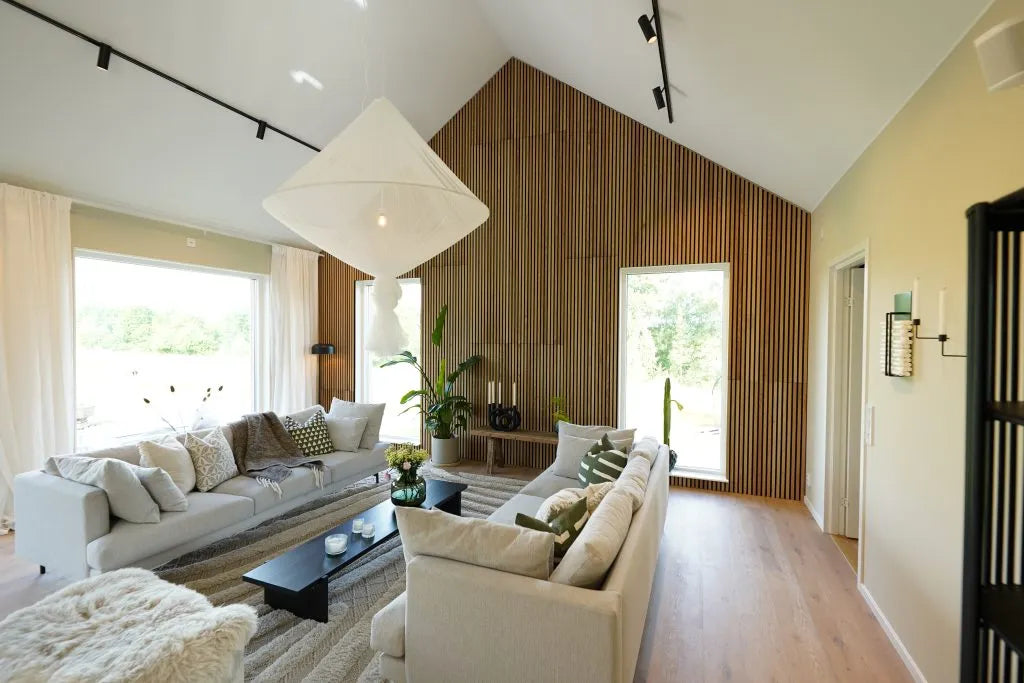 Sala de estar moderna com teto alto, janelas grandes, sofás claros, parede de acento de madeira acústica Akupanel, plantas de interior e luz natural abundante.