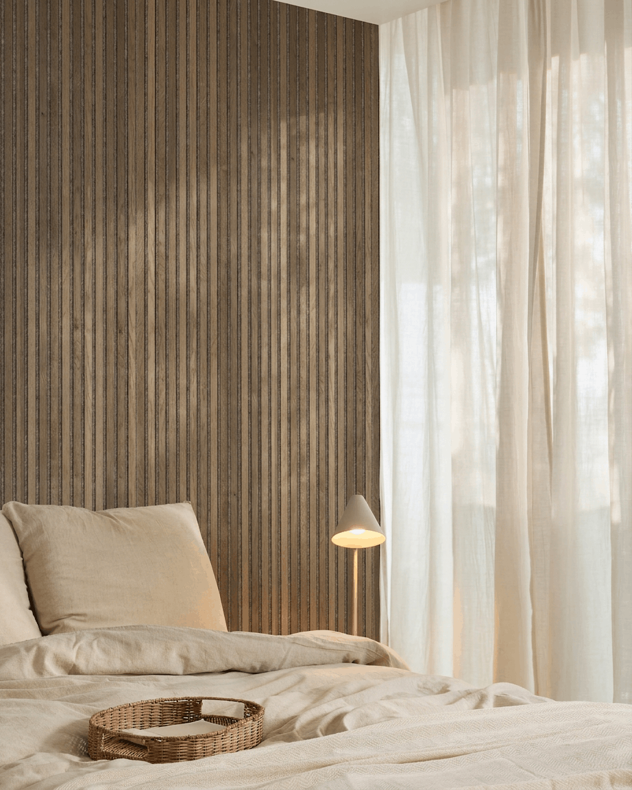 Quarto minimalista com roupa de cama beige, um tabuleiro de verga, painel acústico ripado WoodUpp Akupanel Rustic Grey Oak, um candeeiro branco e cortinas leves.