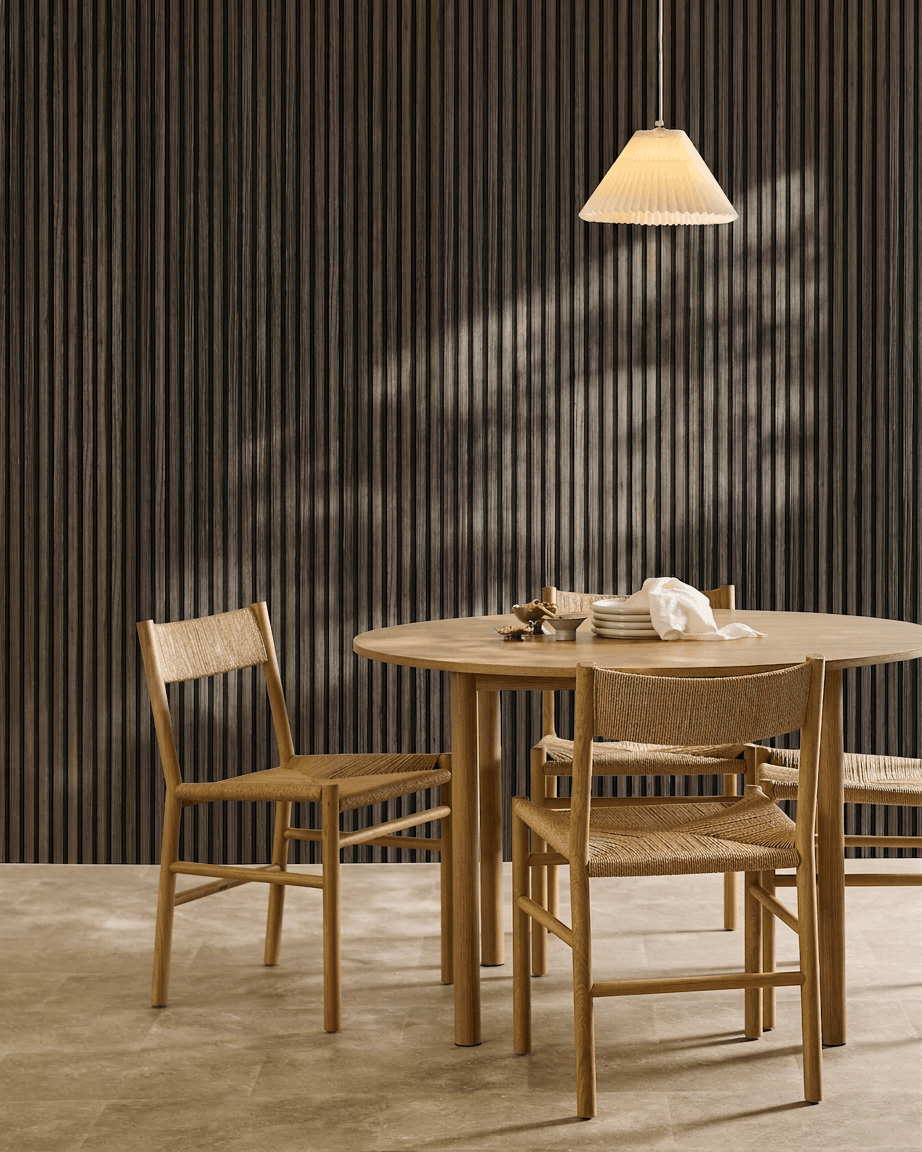 Uma mesa redonda com quatro cadeiras entrançadas está disposta ao lado de um WoodUpp Akupanel Black Oak Painel acústico, sob um candeeiro suspenso plissado.