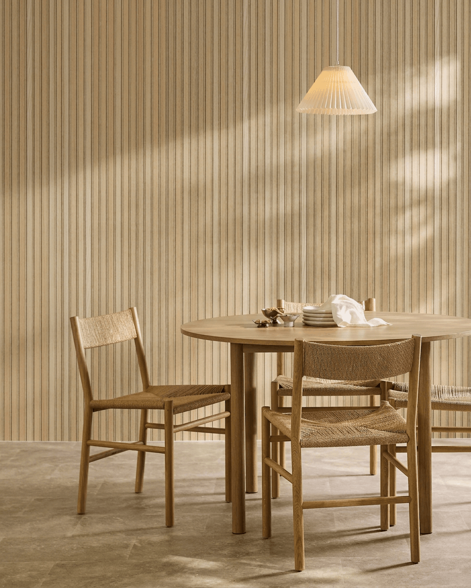 Sala de jantar minimalista com mesa redonda, cadeiras em vime e candeeiro suspenso ao lado de um painel acústico de madeira WoodUpp Akupanel em Carvalho Clássico com feltro beige.