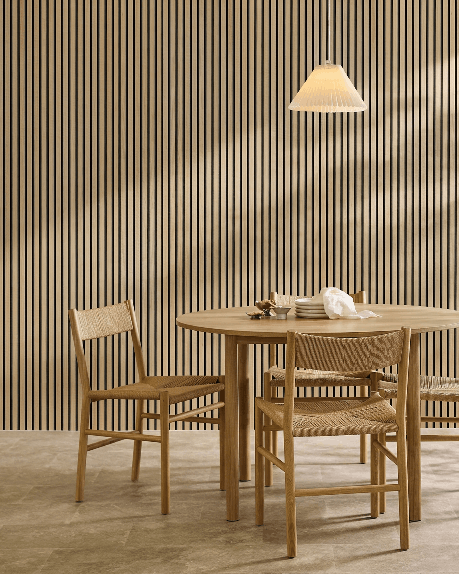 Sala de jantar minimalista com uma mesa redonda, cadeiras em tecido entrançado, candeeiro suspenso e um WoodUpp Akupanel Classic Oak Painel acústico montado na parede.