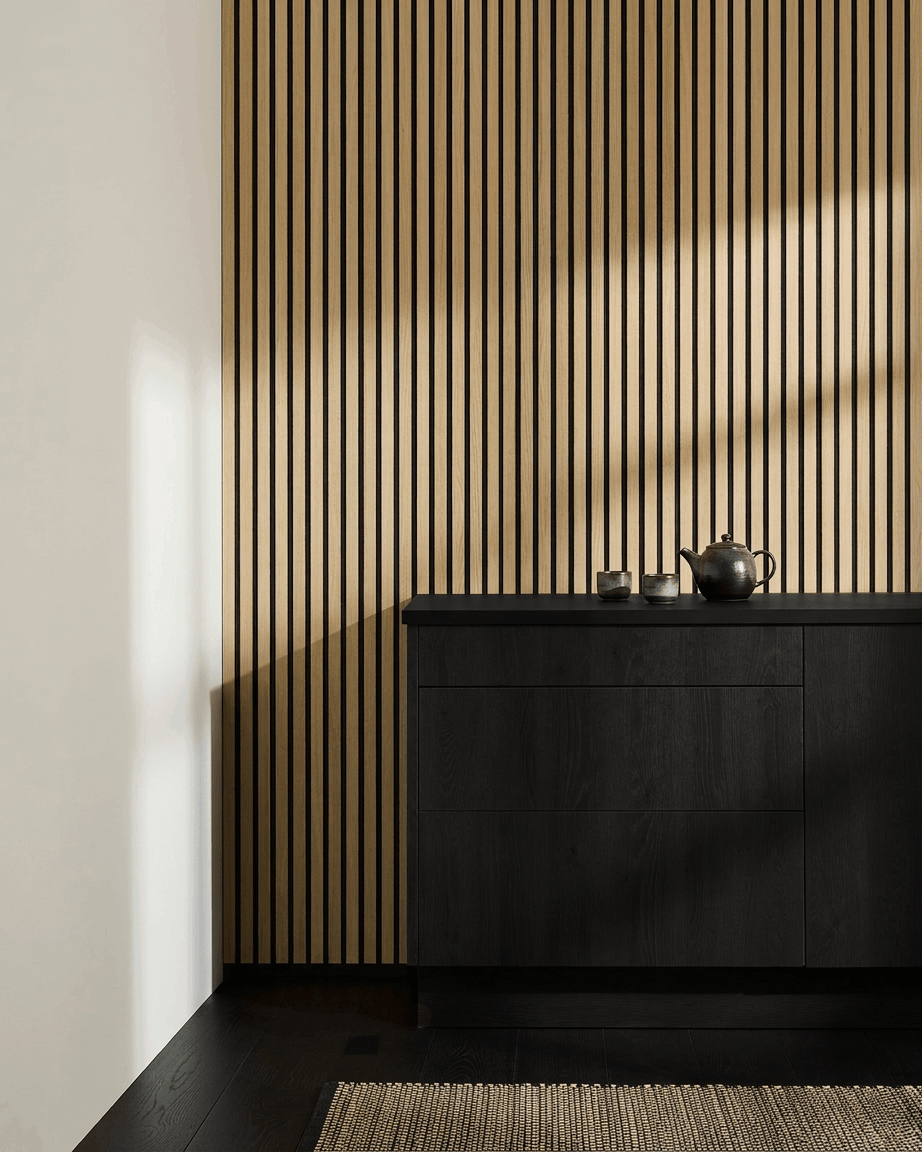 Cozinha nórdica minimalista com WoodUpp Akupanel Painel acústico em Carvalho Clássico, armário black, bule, chávenas e tapete entrançado sobre chão de madeira escura.
