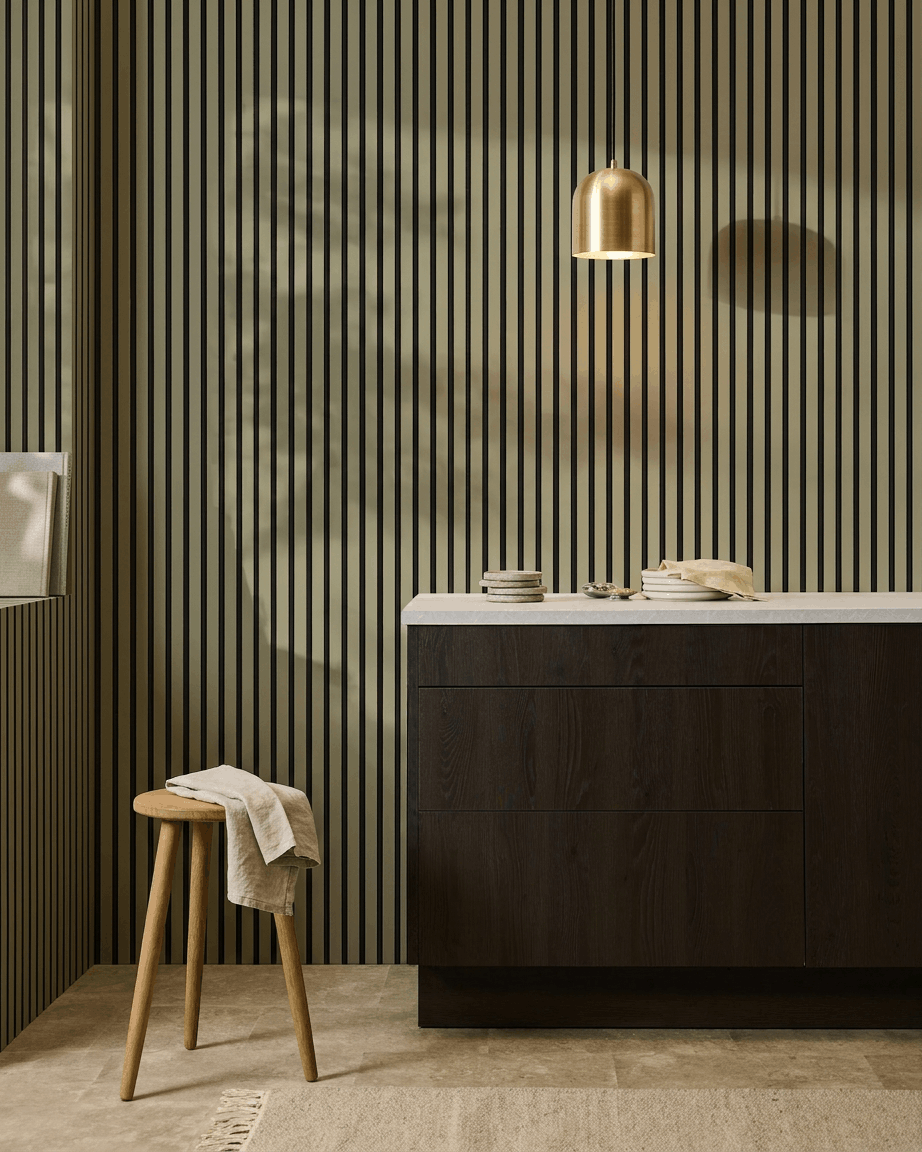 Cozinha moderna com armários de madeira escura, bancada em mármore, candeeiro suspenso brass e um painel acústico ripado WoodUpp Akupanel Colored para absorção sonora.