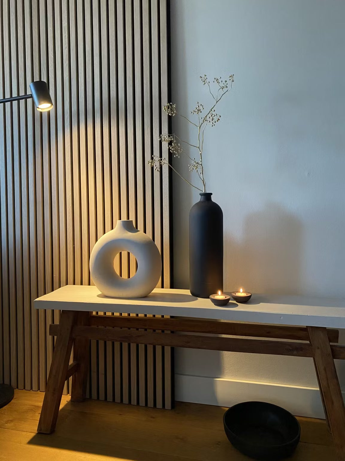 Decoração minimalista com um vaso circular branco, um vaso alto preto com caules secos, duas velas tealight e uma tigela preta em um painel acústico de ripas de madeira Akupanel como pano de fundo.