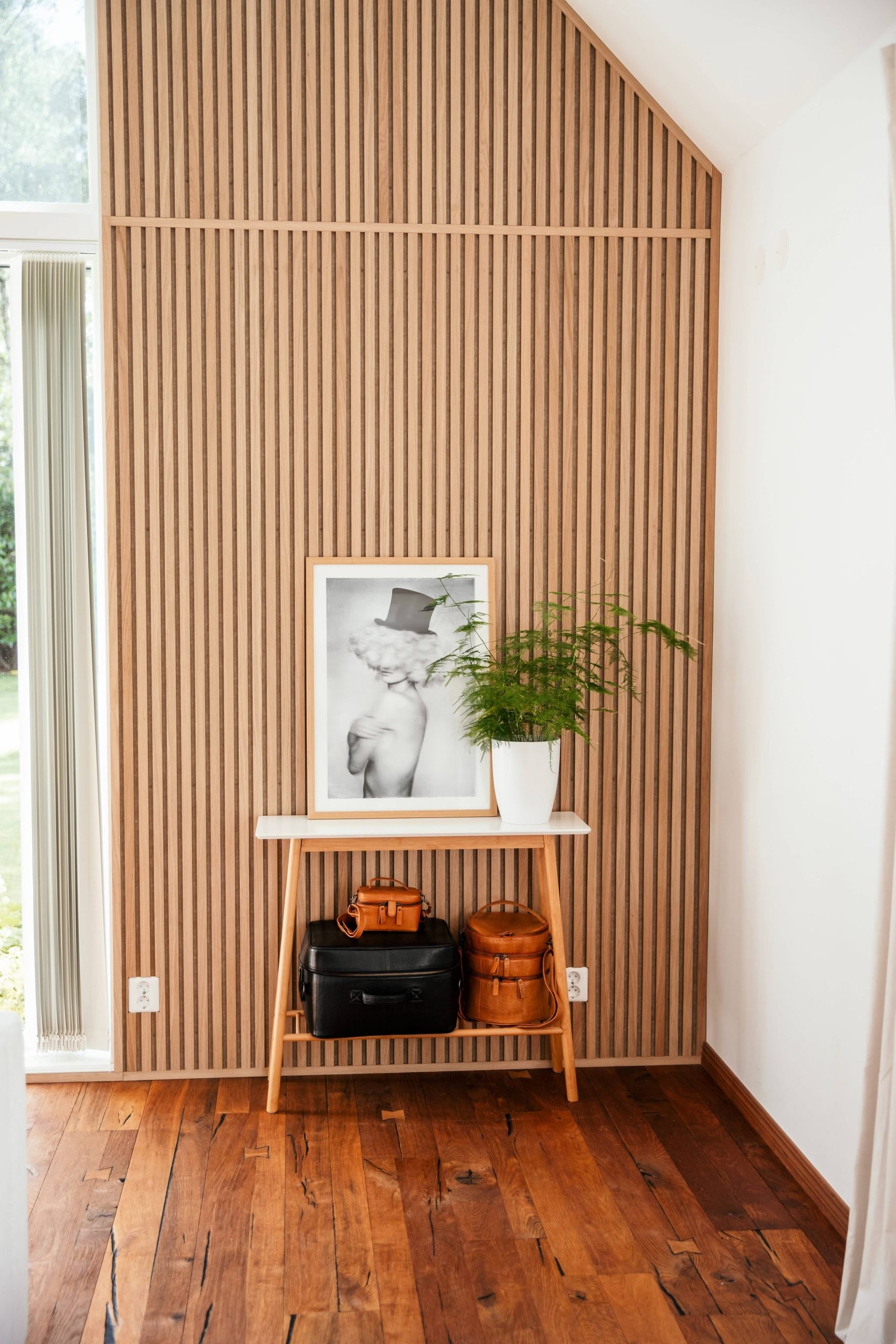 Uma pequena mesa de madeira com sacolas, uma foto emoldurada e um vaso de plantas fica em frente a um painel acústico nórdico de parede de ripas em uma sala iluminada com piso de madeira.