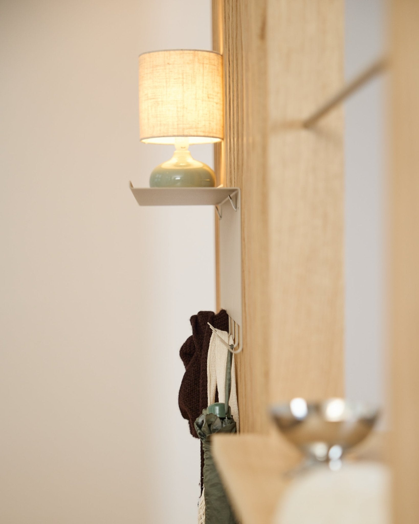 Um cabide Nell da WoodUpp BM1 beige (63,5 x 24 cm) está colocado por baixo de um candeeiro de mesa small, junto a uma parede em madeira nórdica clara Painel acústico.
