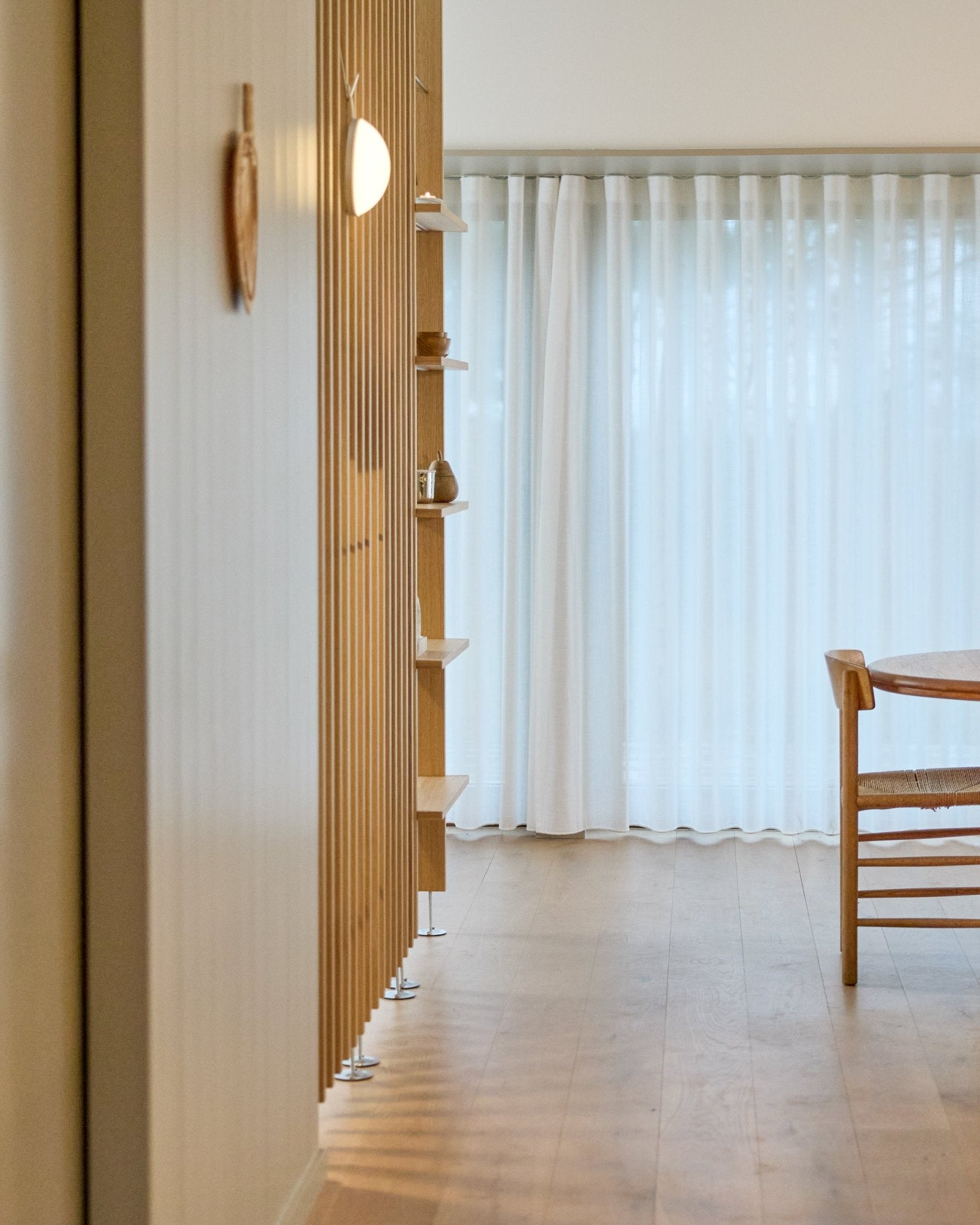 Espaço minimalista com uma divisória de espaços WoodUpp Embrace, do chão ao teto, chão de madeira, prateleiras, cadeira e cortinas leves sobre janelas large.