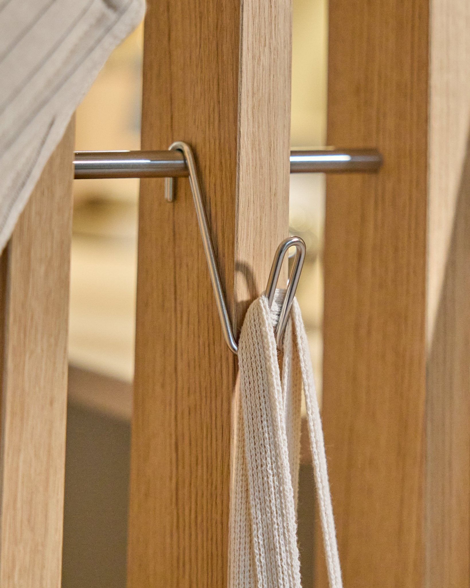 O Alva Wire Hook / BM1 (aço inoxidável Steel, curto) da WoodUpp está montado numa calha de madeira Painel acústico, segurando uma correia branca com haste metálica; fundo beige.
