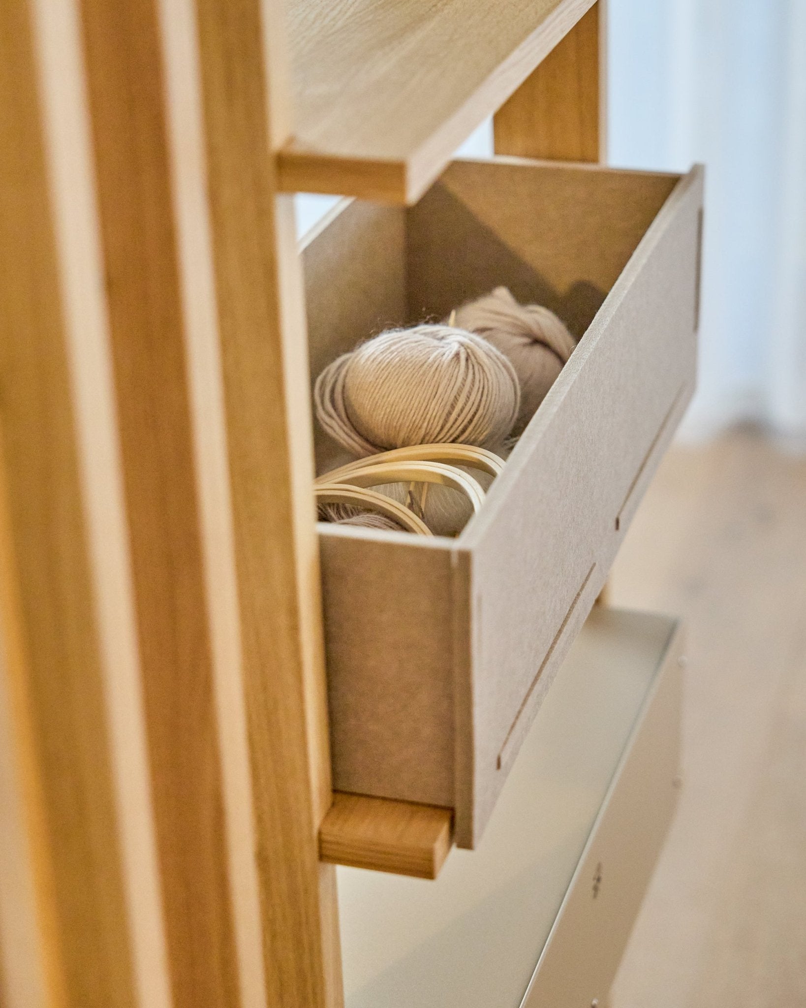 A WoodUpp Vigg Felt Box / BM2 em beige guarda novelos de lã leves e bastidores de bordado em madeira numa estante de madeira, complementando painéis acústicos nórdicos.