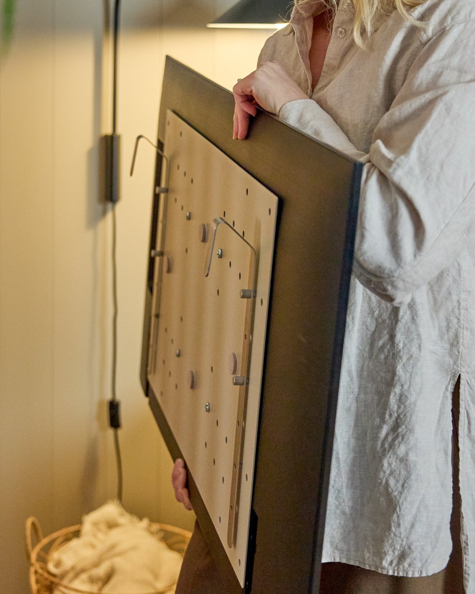 Uma pessoa com uma camisa clara segura o WoodUpp Nell BM1 TV-Mount em beige, 64x44 cm — compatível com painéis acústicos de madeira nórdicos, para instalações sem parede.