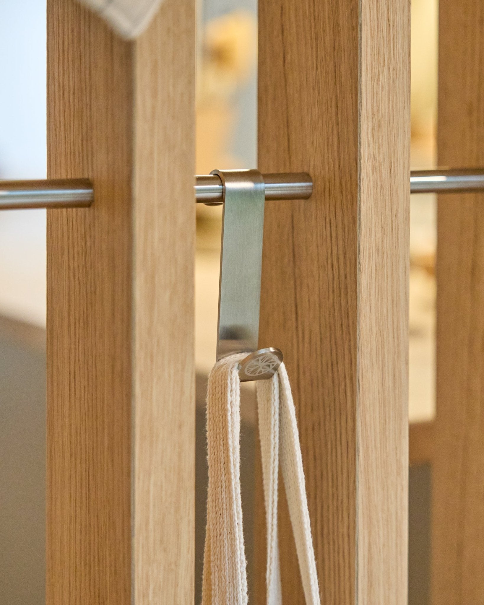 Um gancho Alva S-Hook da WoodUpp em steel inoxidável fixa uma correia beige a uma haste montada entre painéis acústicos de ripas de madeira vertical. O conjunto inclui 2 ganchos.