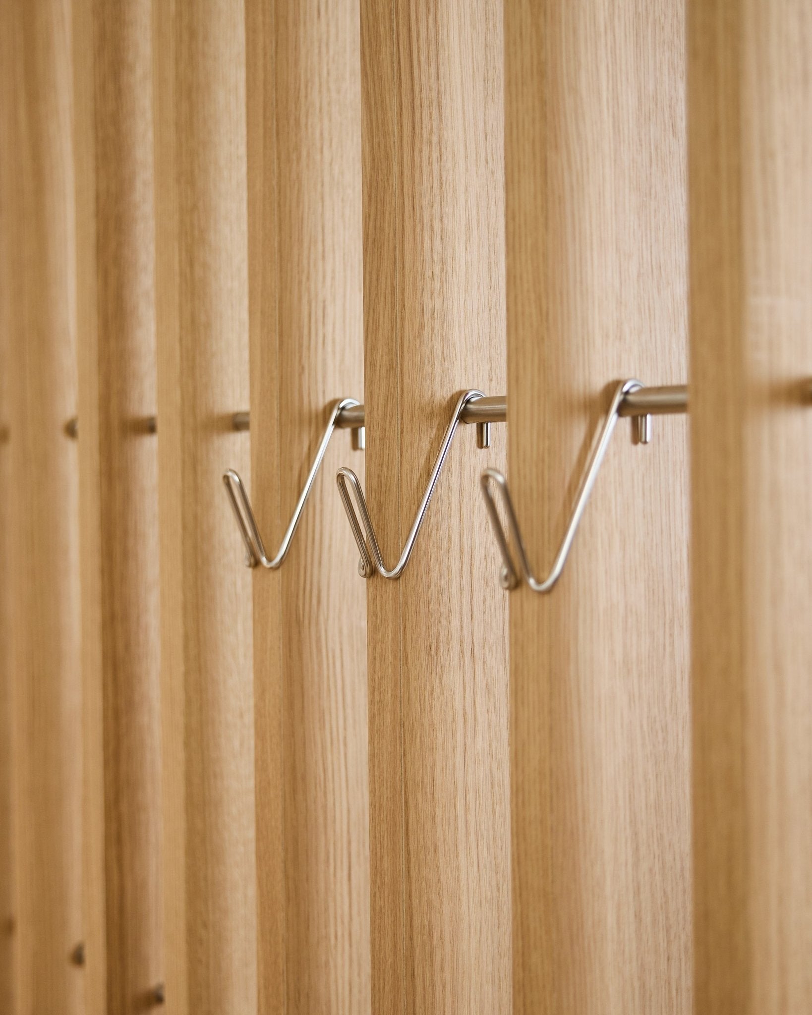 Três acessórios Alva Wire Hook / BM1 curtos em aço inoxidável steel da WoodUpp estão montados de forma uniforme em fila num painel acústico ripado de madeira vertical.
