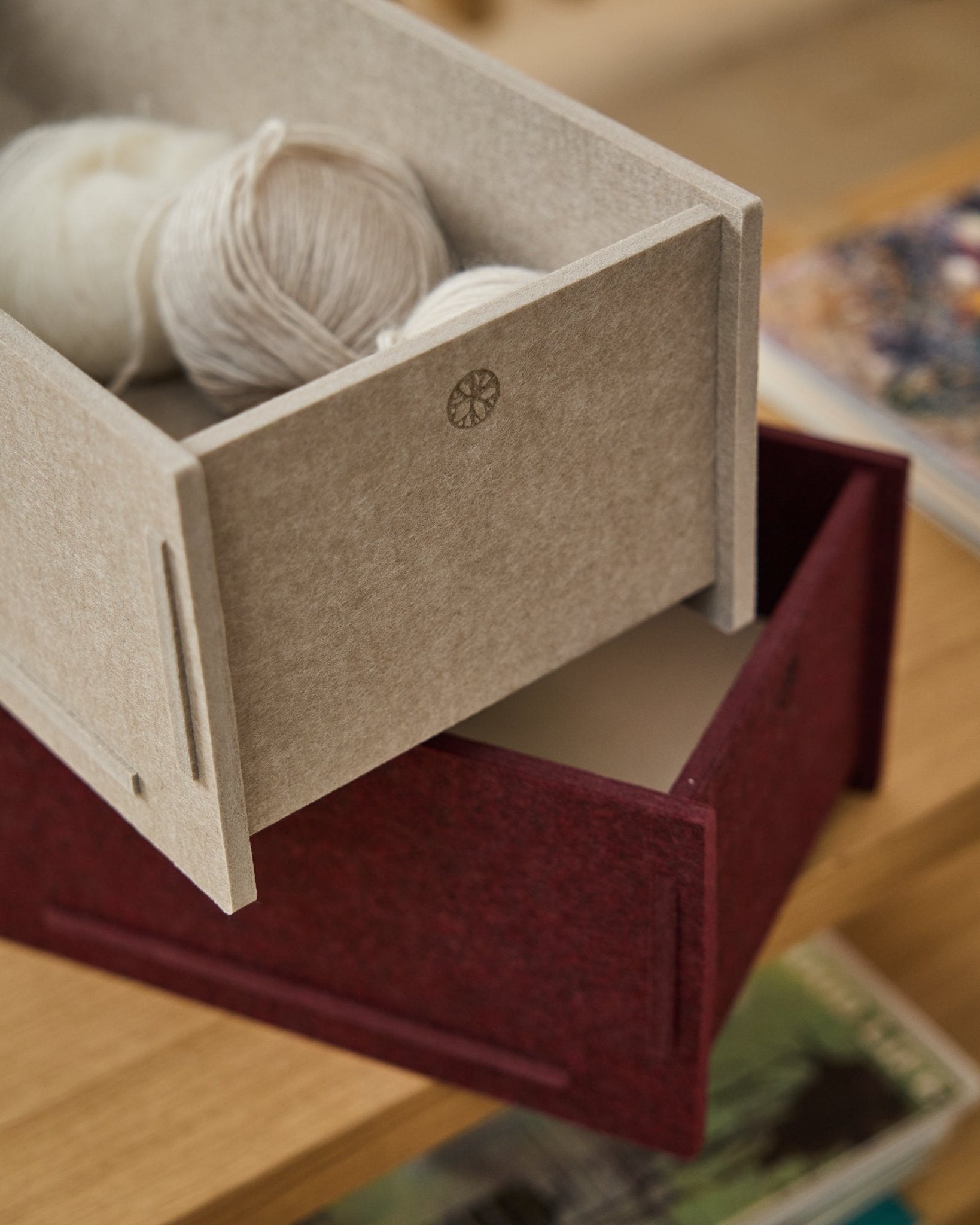 Uma caixa de arrumação em feltro PET beige, cheia de fios, está sobre uma Vigg Felt Box/BM2 fechada da WoodUpp em ox red, assente num soalho de madeira, com livros em segundo plano.