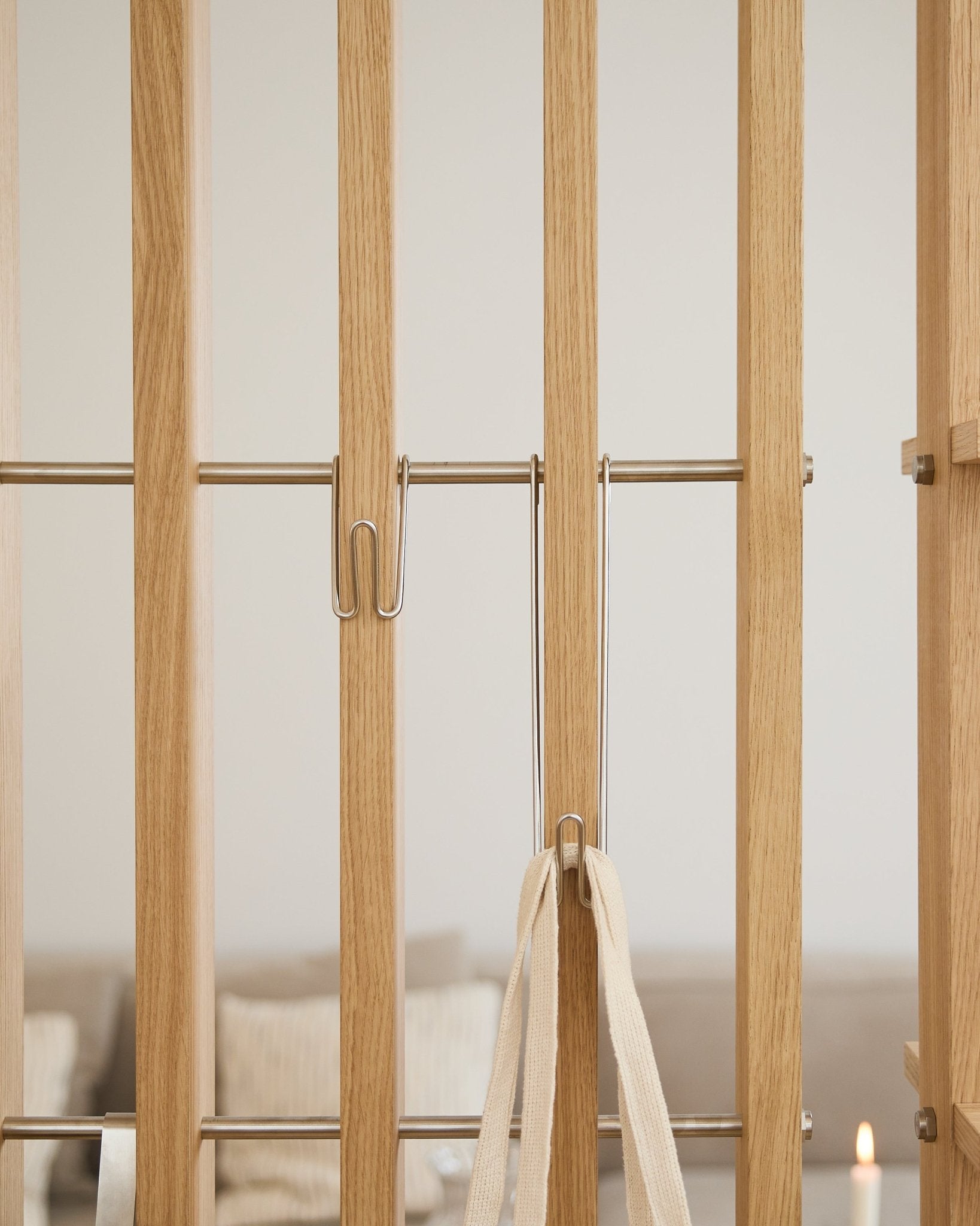 Grande plano do WoodUpp Alva | Wire Hook BM1 (aço inoxidável steel, comprido, conjunto de 3) preso a ripas em Painel acústico claro com uma correia branca; sofá e vela desfocados ao fundo.