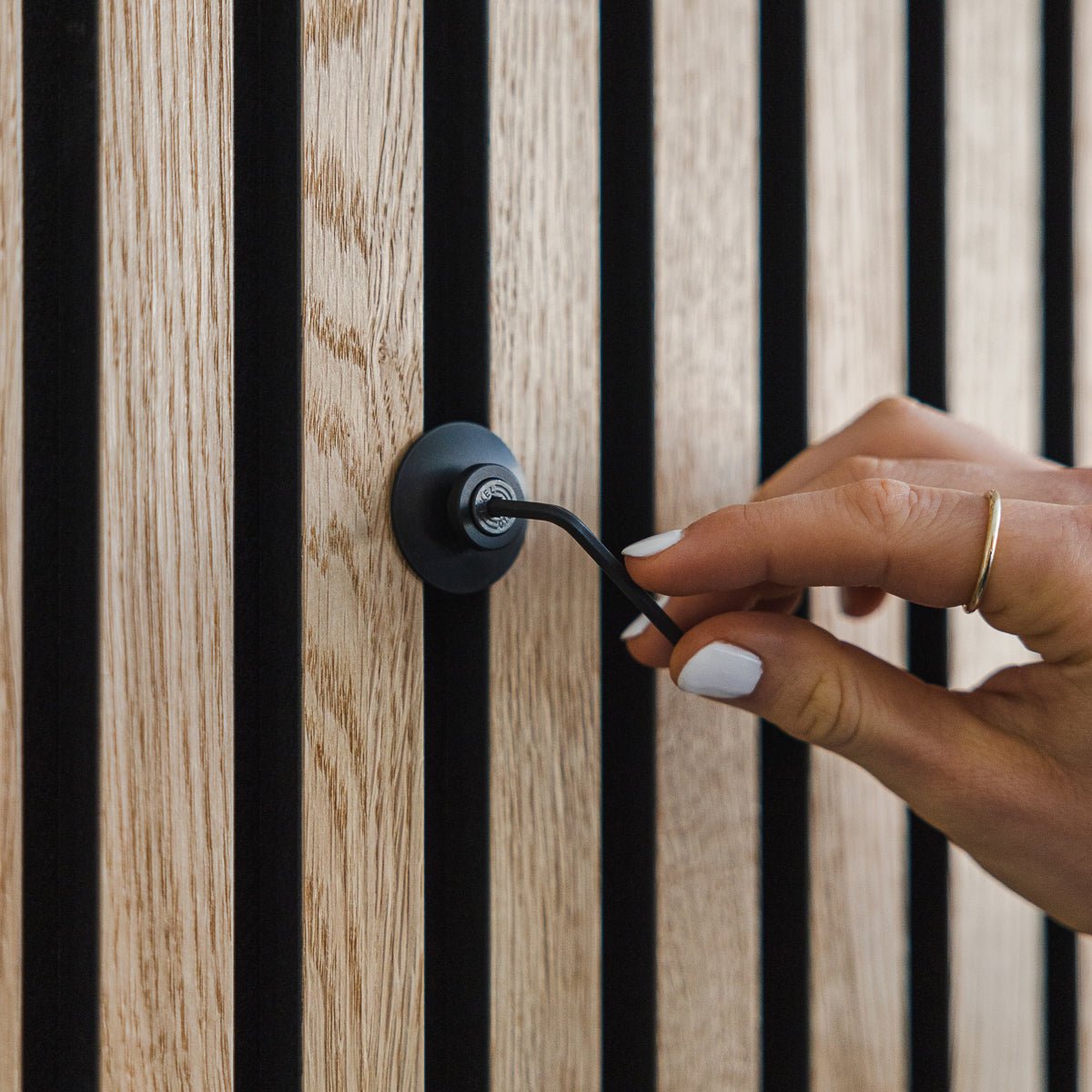 Uma mão com verniz branco nas unhas roda um gancho de alumínio black | Heavy Dupps da WoodUpp num painel acústico de madeira vertical Akupanel.