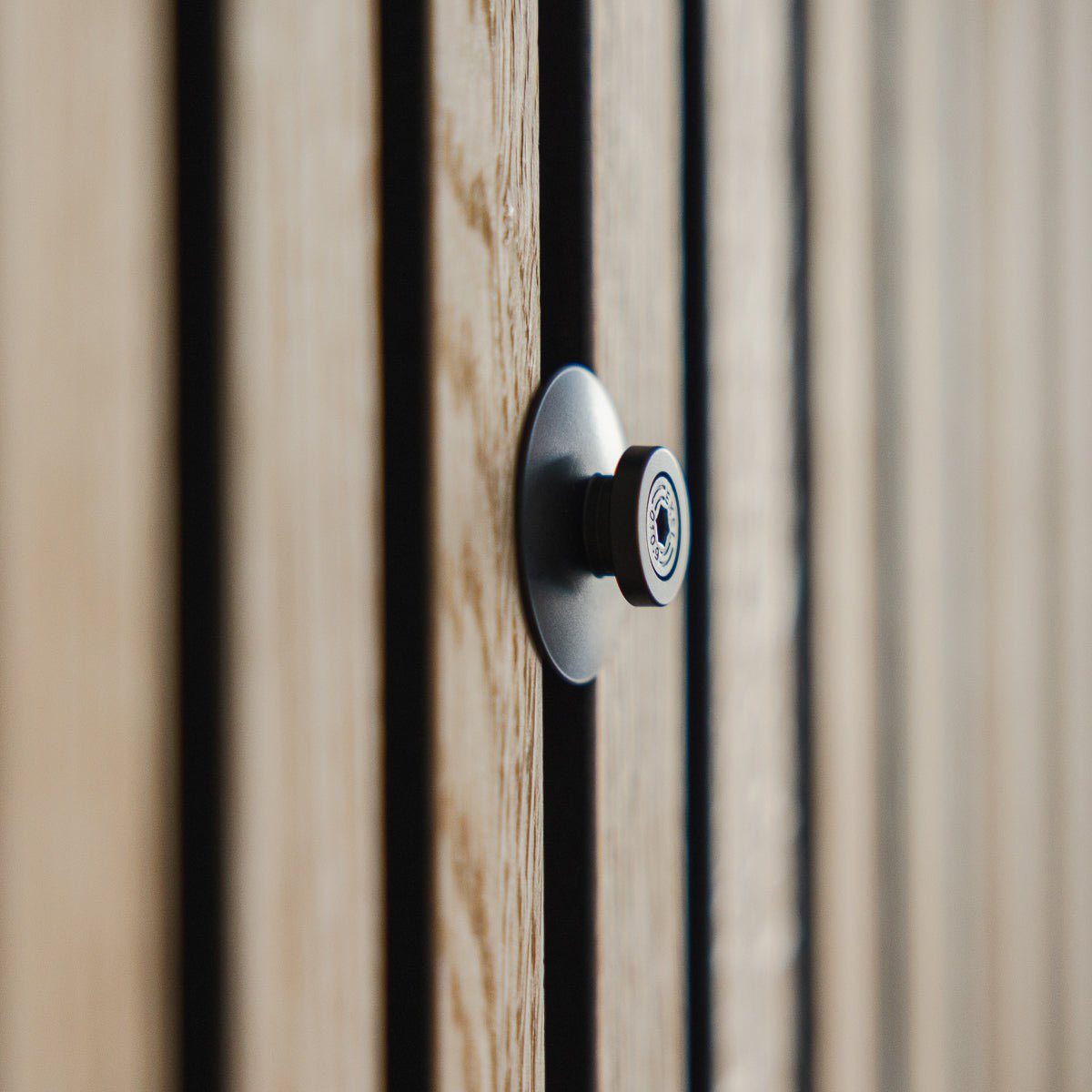 Grande plano do Dupps | Óculo Heavy da WoodUpp em alumínio black, instalado numa porta de madeira com ranhuras e ripas acústicas Akupanel, com a lente em foco.