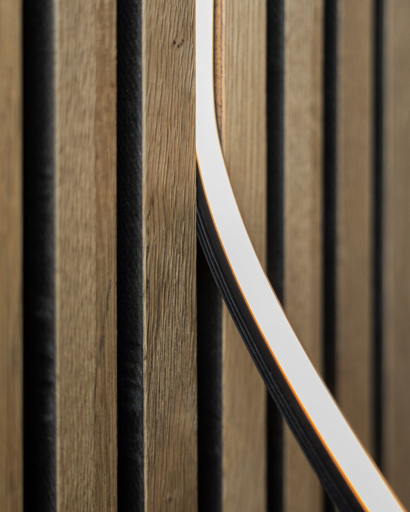 Close-up of WoodUpp Akupanel acoustic panel in natural wood finish with black grooves and a 300 cm Lumi LED striplight, forming a modern, linear pattern.