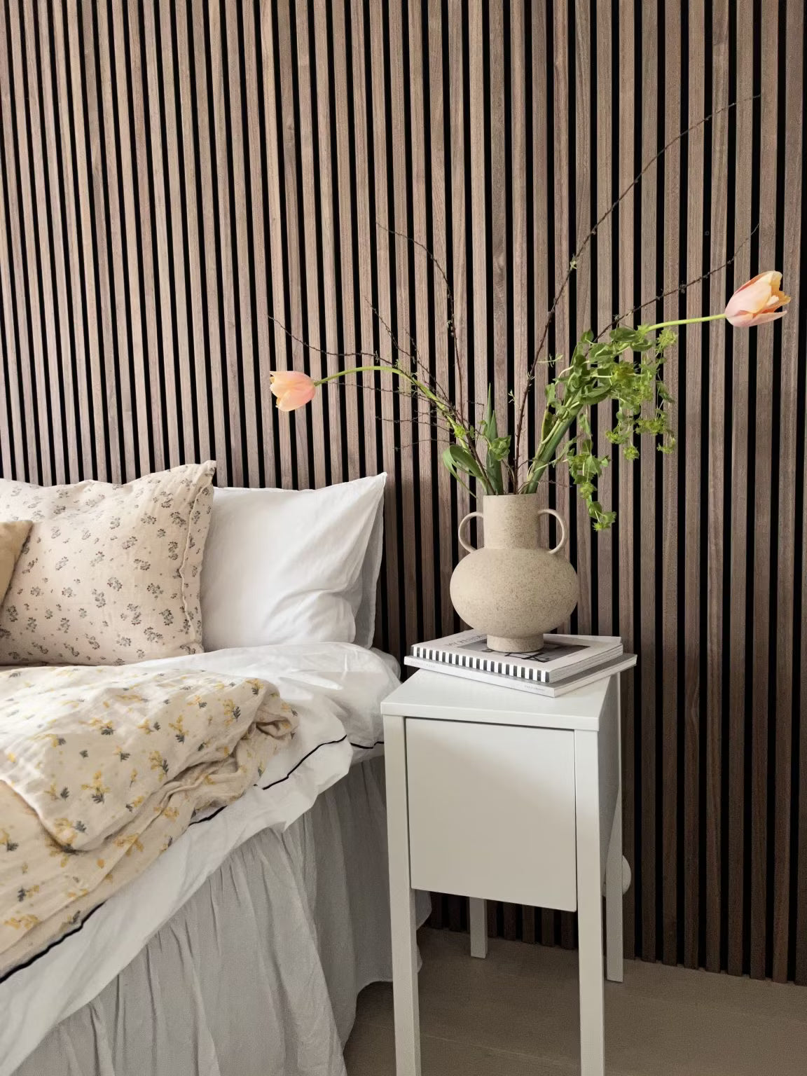 Um quarto com uma cama branca com roupa de cama floral e um vaso de flores, com uma parede nórdica de ripas acústicas de madeira Akupanel atrás da cama.
