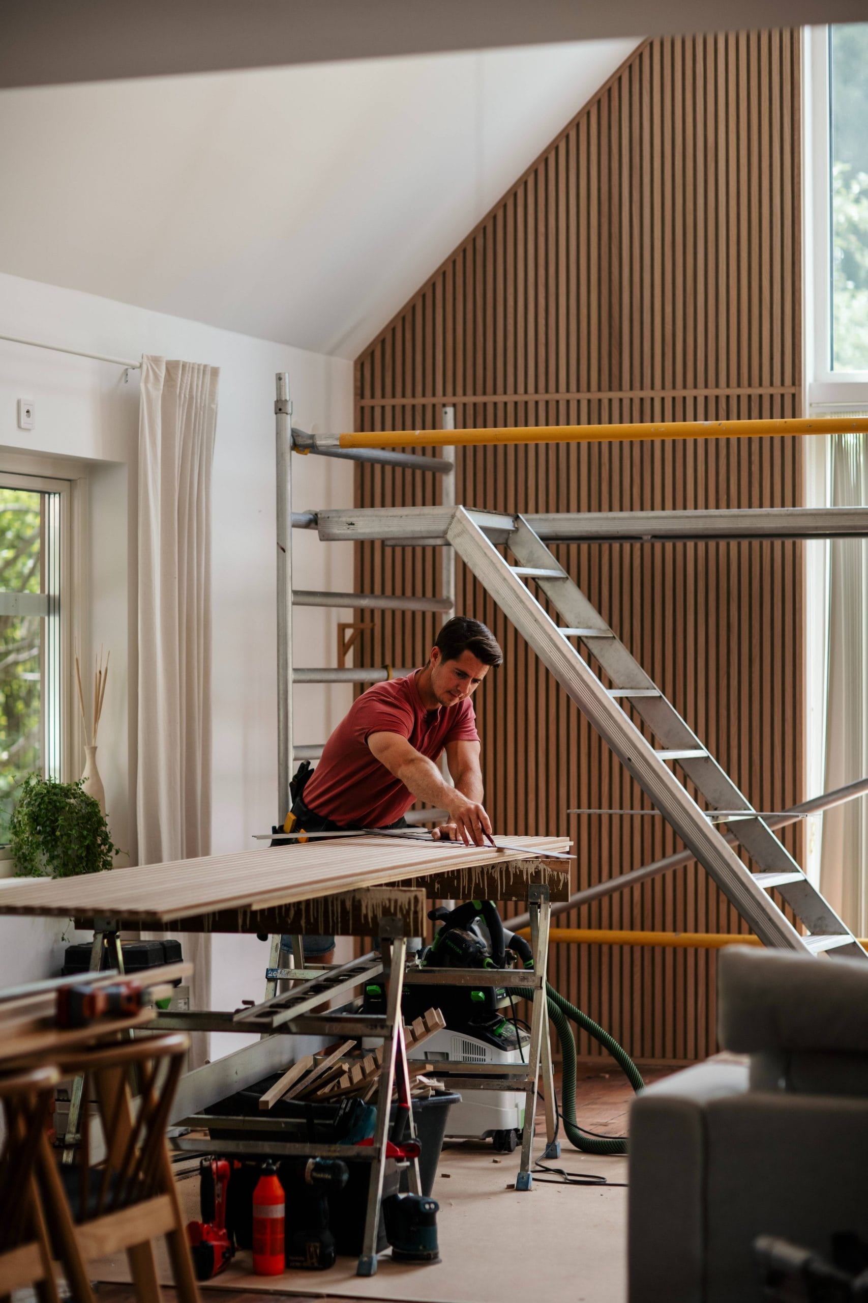 Um homem mede uma prancha de madeira em uma bancada de trabalho em uma sala moderna, parcialmente reformada, com andaimes e grandes janelas, sugerindo a instalação de um painel acústico.