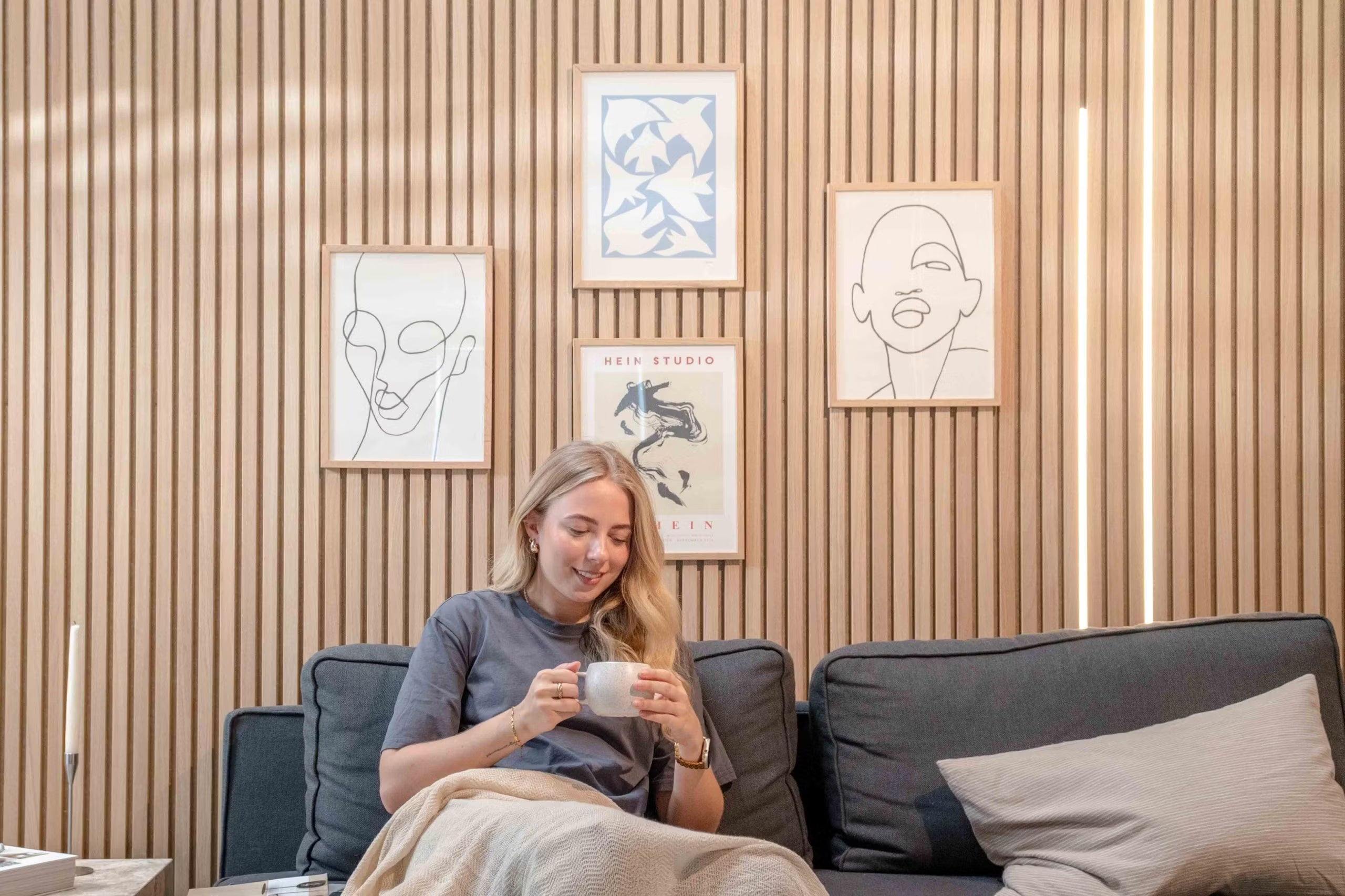 Mulher relaxando em um sofá com uma caneca, sorrindo sob arte de linha minimalista e decoração abstrata montada em painéis de parede de ripas acústicas nórdicas texturizadas.