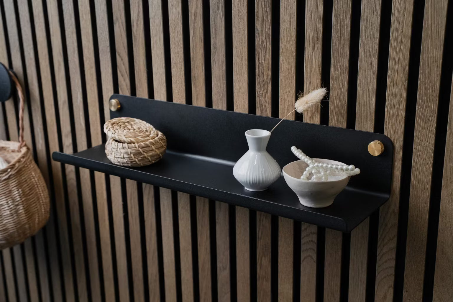 Uma prateleira de parede preta com uma pequena cesta de vime, um vaso branco e uma tigela de pérolas de cerâmica montada em um painel de parede de ripas acústicas de madeira nórdica.