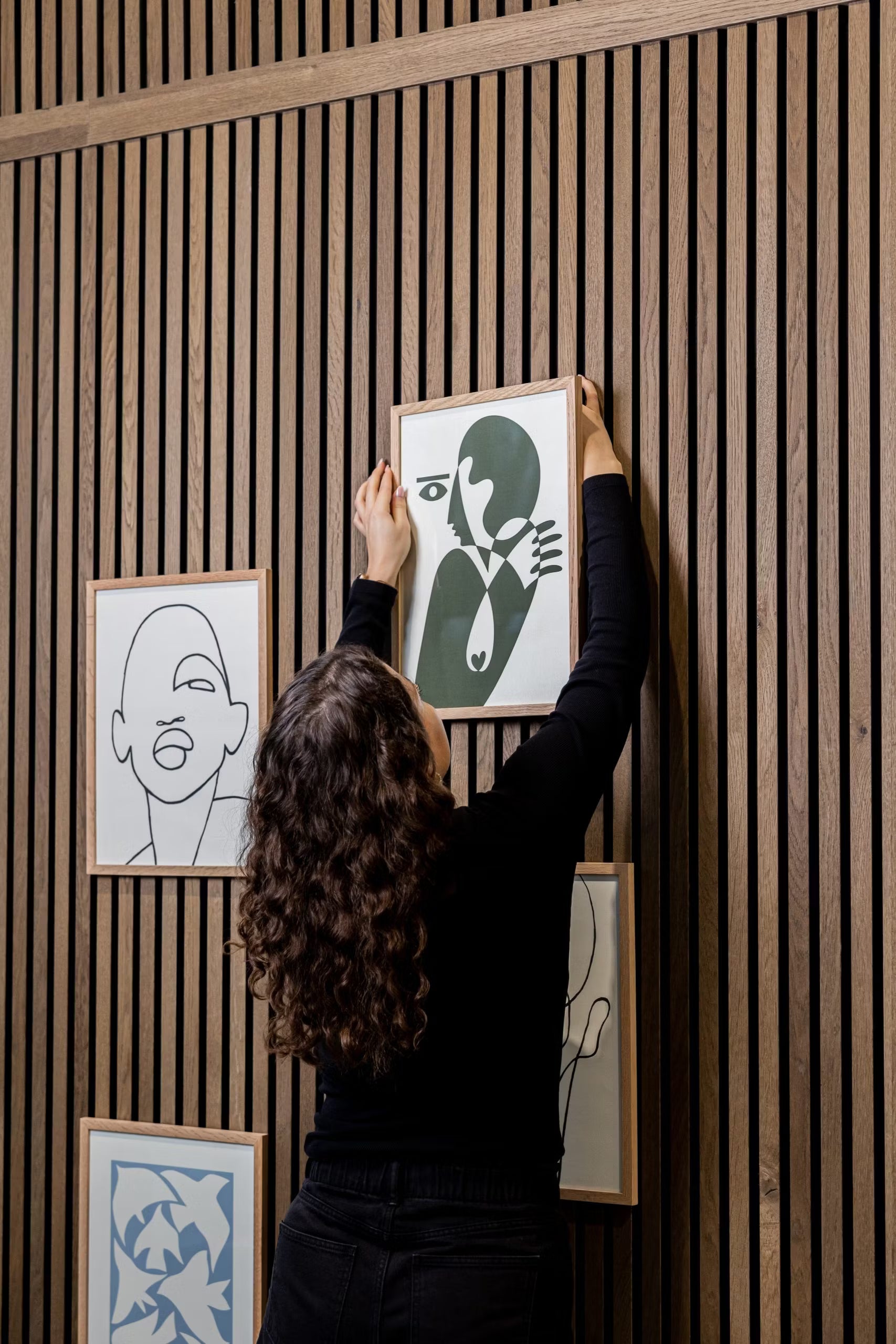 Uma pessoa de cabelo encaracolado pendura um acessório WoodUpp® CREATE DUPPs num painel acústico ripado de madeira, rodeada por arte minimalista e decoração moderna.