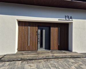 Entrada de casa com painéis verticais de ripas de madeira WoodUpp em acabamento natural cobrindo a parede, duas luzes embutidas verticais LED WoodUpp, porta central cinza e placa do número 13A em metal WoodUpp, soleira e pavimento em pedras rústicas em frente.