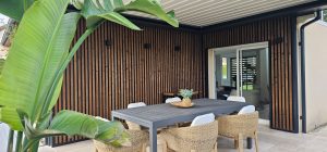 Varanda com revestimento ripado vertical WoodUpp: painéis de madeira escura em lâminas regulares montadas com perfis pretos, criando parede texturizada. Mesa de jantar cinza e cadeiras de fibras naturais à frente; planta tropical à esquerda, porta de correr envidraçada que abre para o interior.